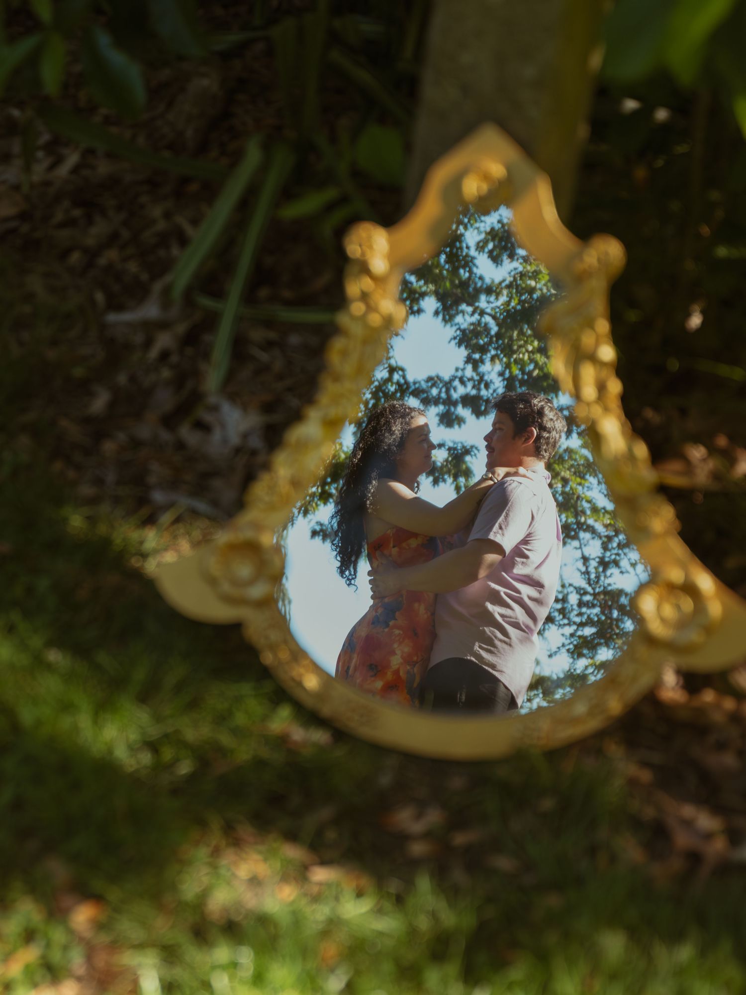 A couple embraces reflected in an ornate gold mirror placed outdoors among lush green trees and grass.