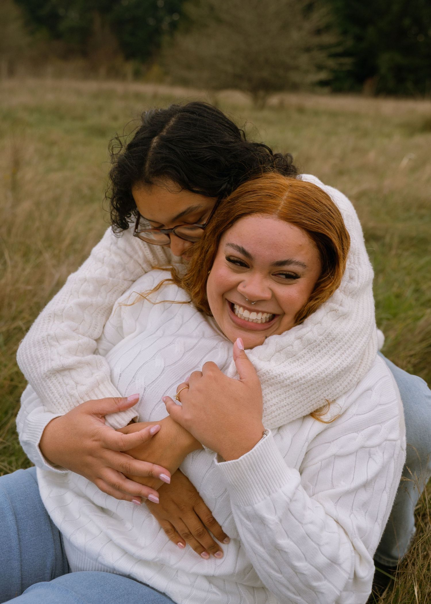 Two women in matching white cable-knit sweaters share a warm embrace and laugh together in an outdoor field setting.