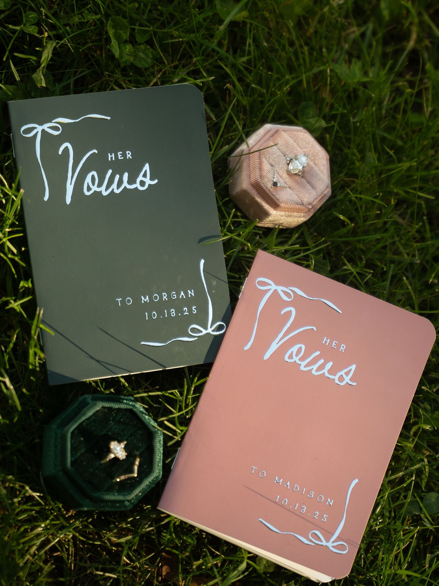 Two Her Vows booklets in green and pink with wedding rings in velvet boxes on green grass, dated 10.18.25.