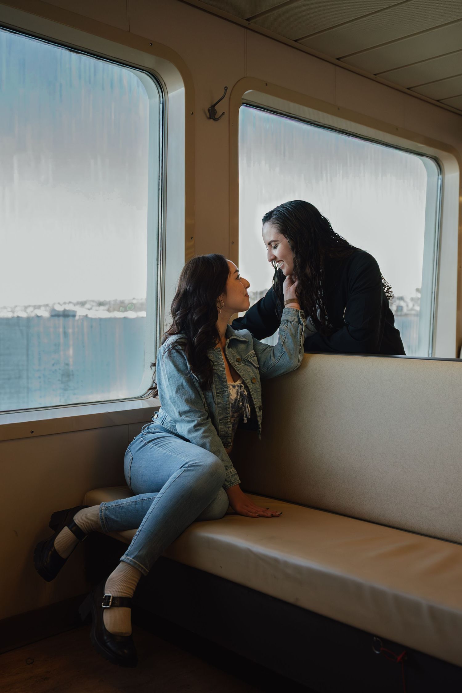 Two women share a joyful moment sitting by rain-streaked windows on a ferry boat, smiling at each other.