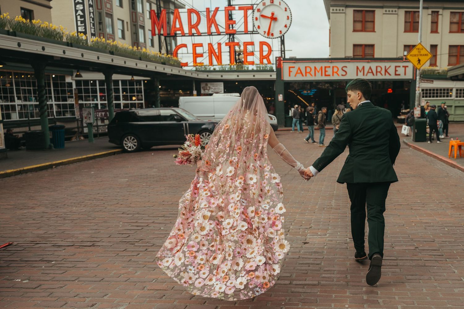 Bride in floral gown and groom in green suit hold hands at Pike Place Market Farmers Market in Seattle.