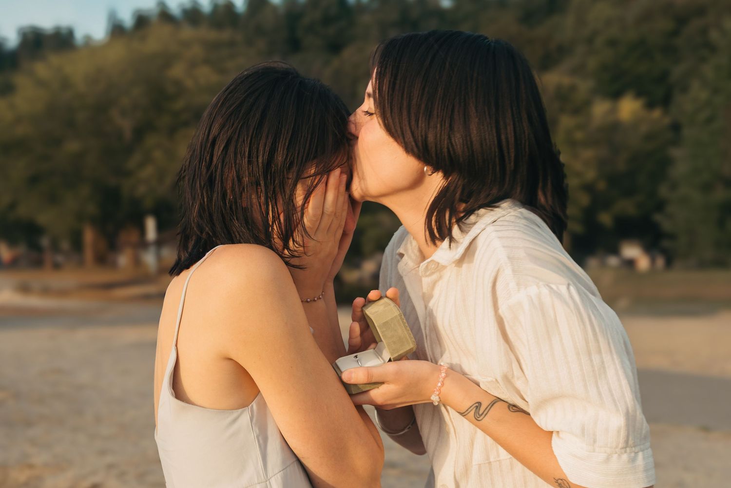 Two people share an intimate moment on a beach at sunset with golden light filtering through the trees.