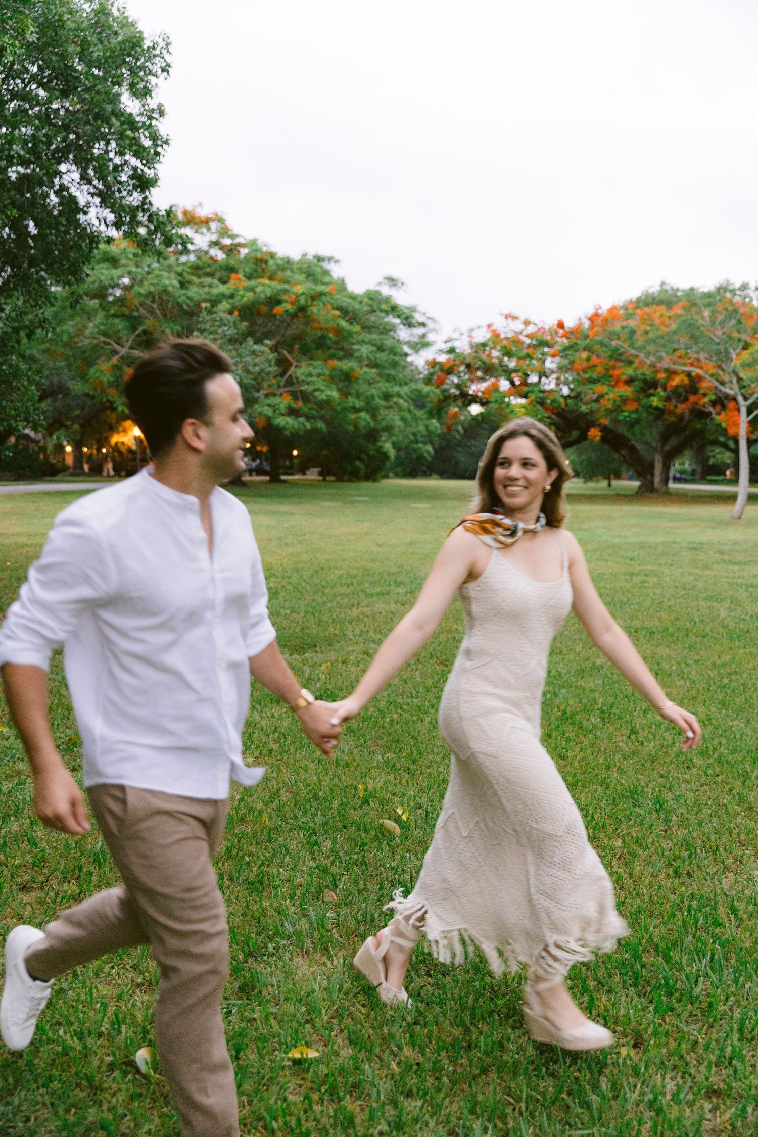 A joyful couple holds hands while running through a lush green park, dressed in white and beige outfits.
