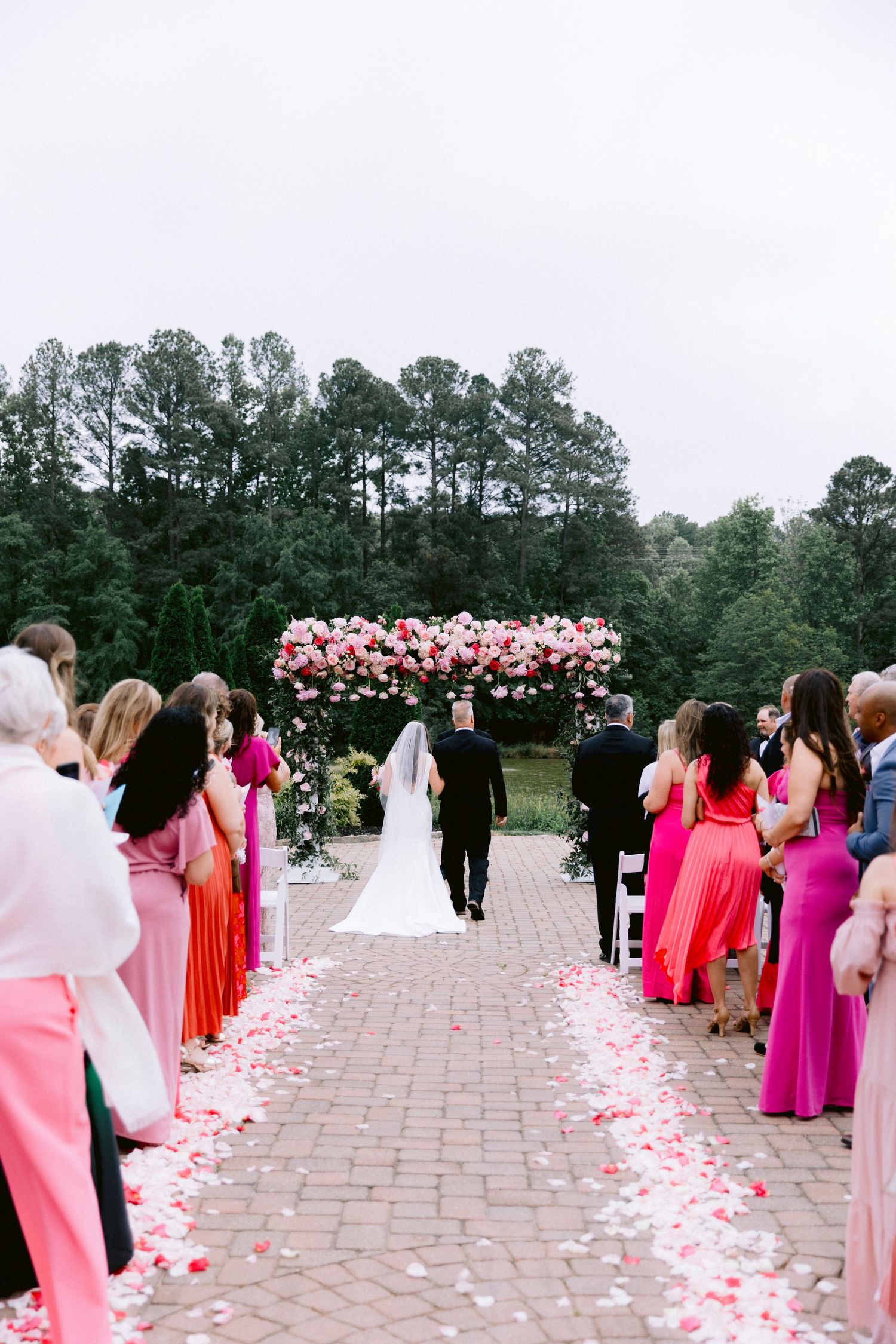 Bride walks down petal-lined aisle toward floral arch, guests in pink and red attire at outdoor wedding ceremony.