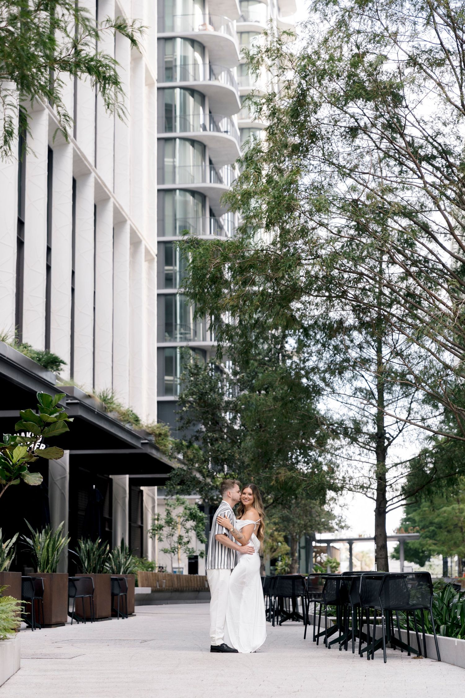 Couple embracing outdoors near a modern white high-rise building surrounded by lush greenery and black outdoor furniture.