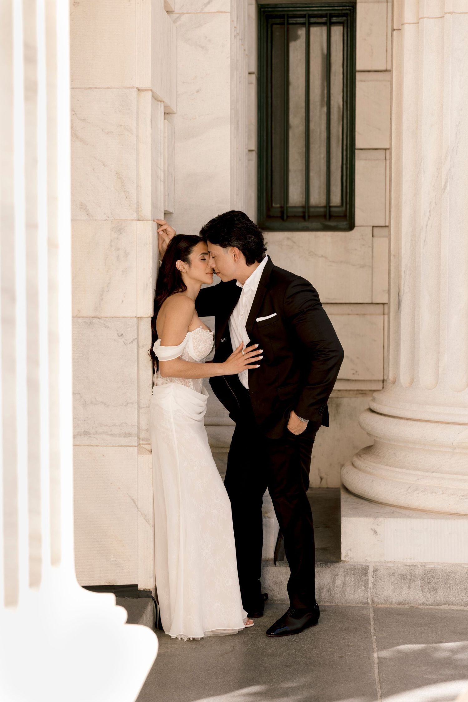 Bride and groom sharing a romantic kiss between marble columns in elegant wedding attire.
