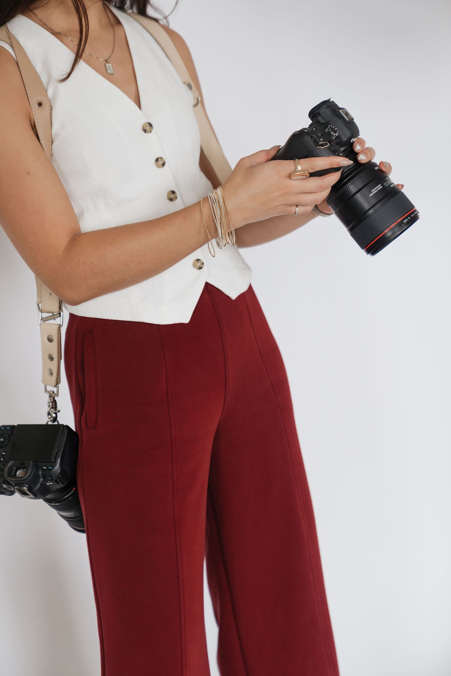 Woman in white vest top and red wide-leg trousers holding a DSLR camera with a black bag.