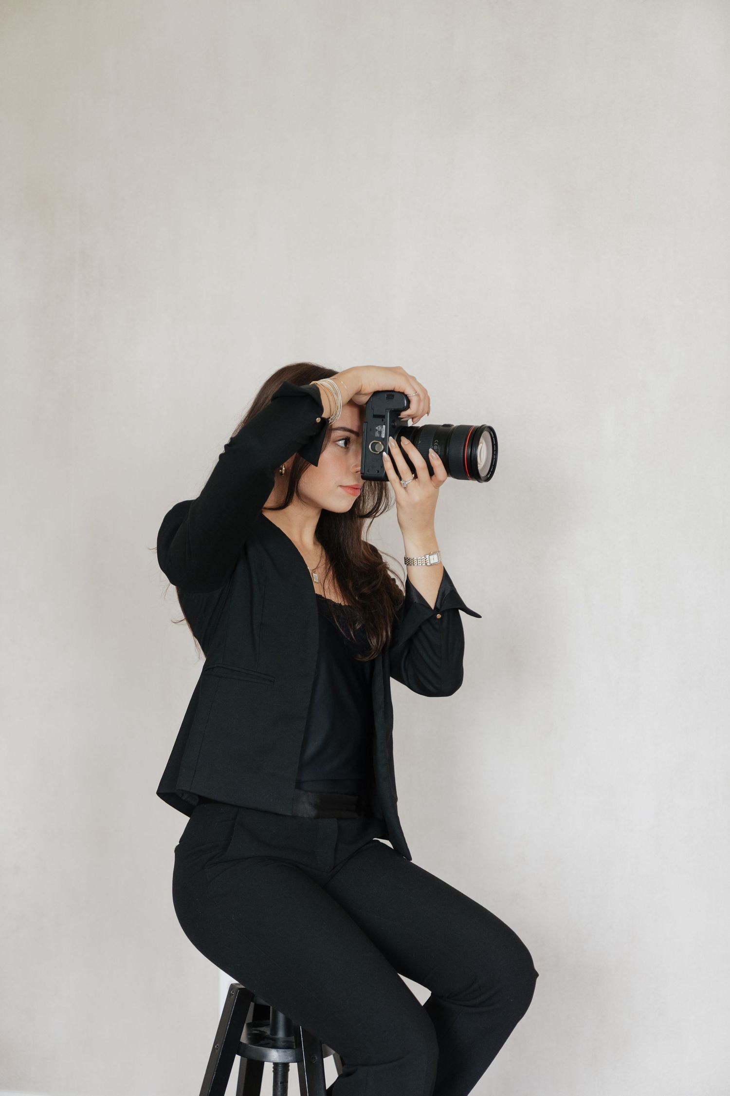 A professional female photographer in a black suit sits on a stool, holding a DSLR camera up to take a photo.