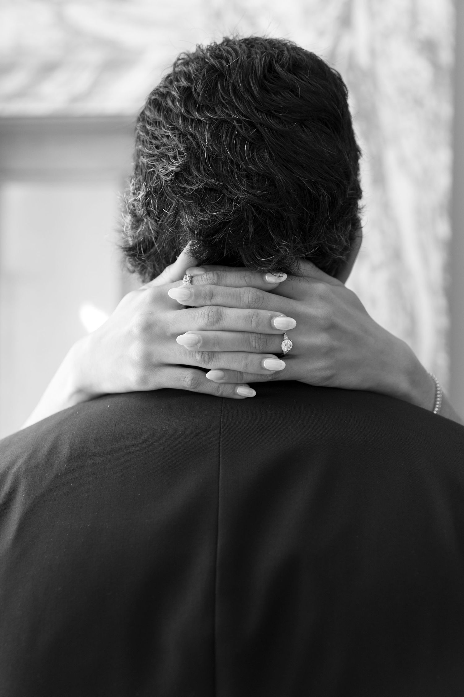 Black and white photo of a woman's hand with engagement ring resting on a man's neck in a romantic embrace.