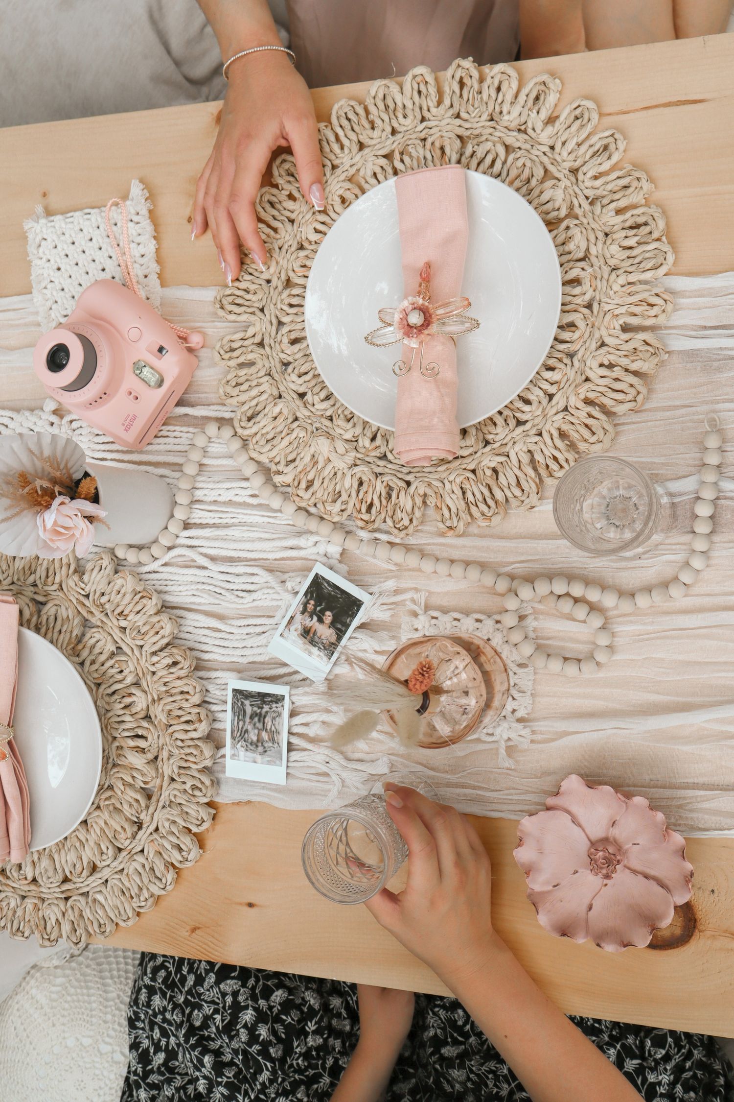 Overhead view of a table setting with macrame placemats, pink decor, and vintage polaroid photos on a wooden surface.