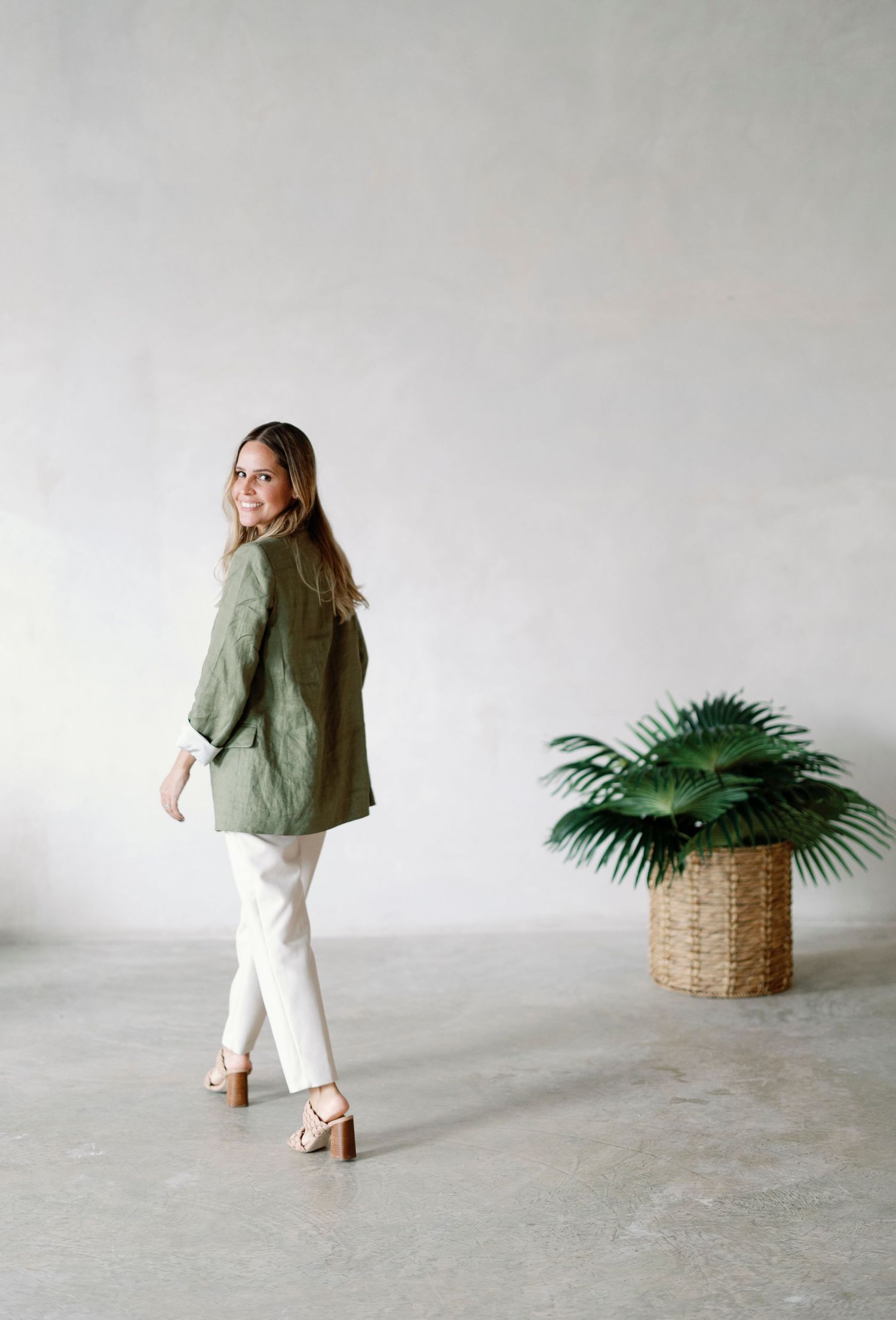 A woman in a green coat stands in a room with a potted plant.