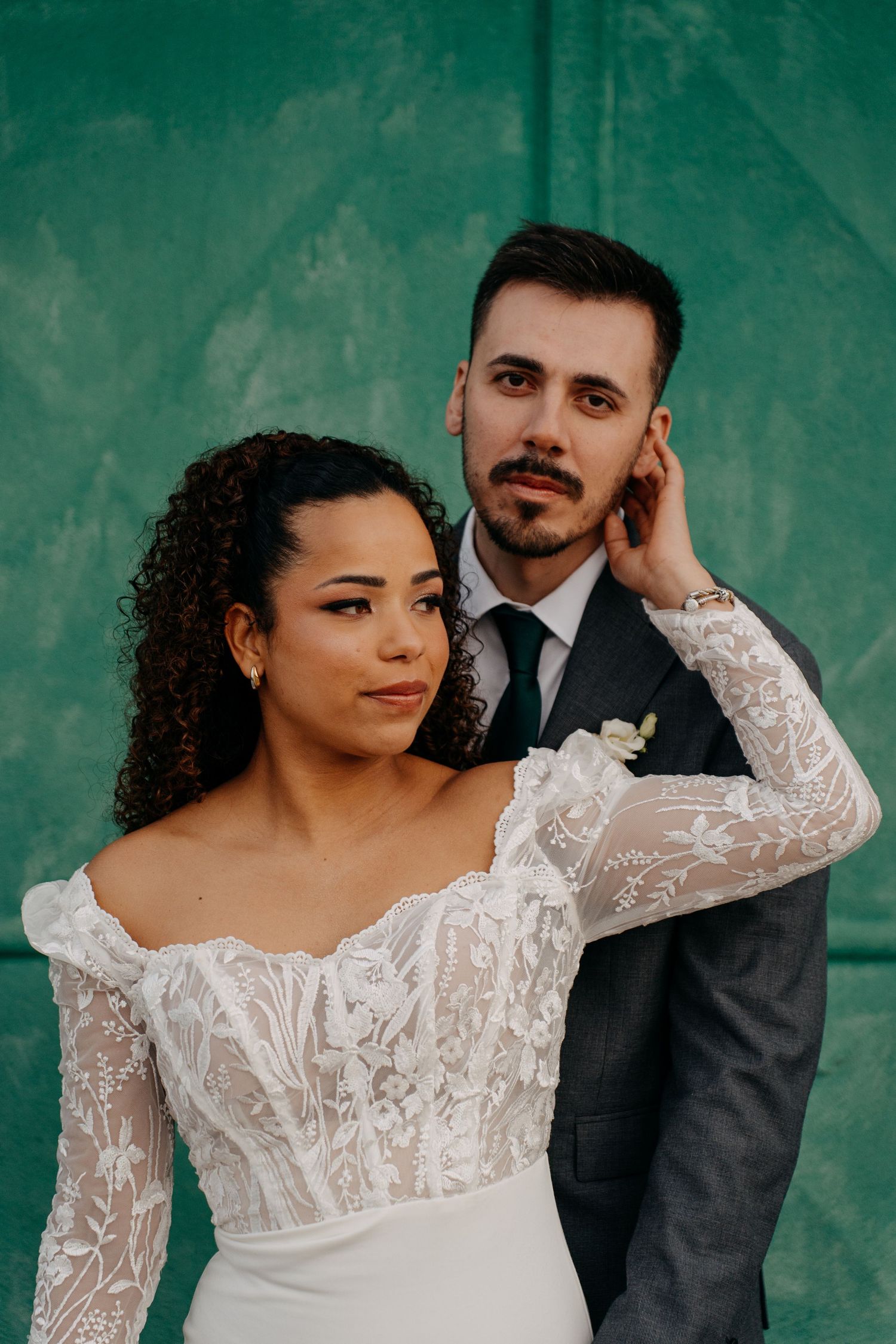 A wedding couple in formal attire pose intimately against a teal wall.