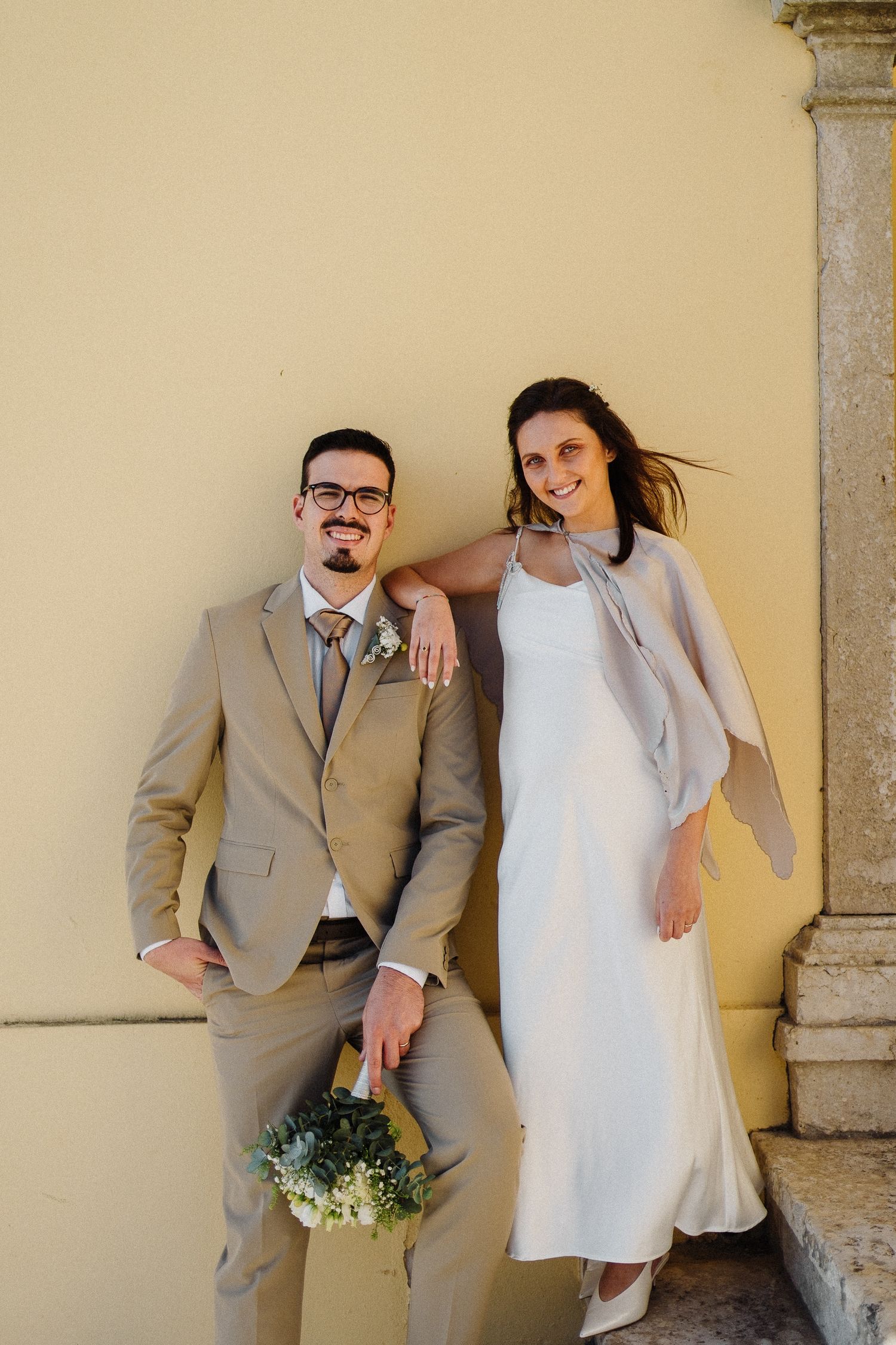 A wedding couple in coordinating beige and white attire pose against a yellow wall with bouquet.