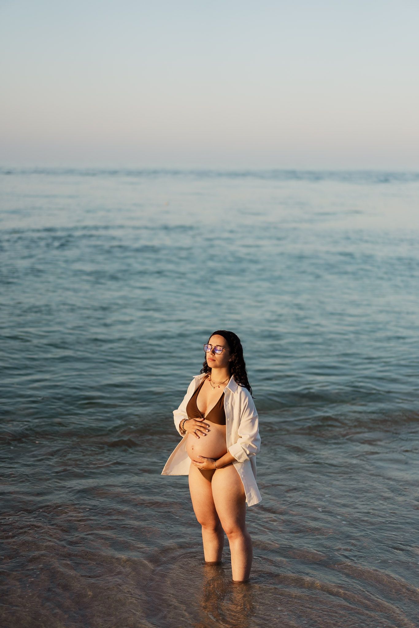 Person in swimwear and open shirt standing in shallow ocean water at sunset.