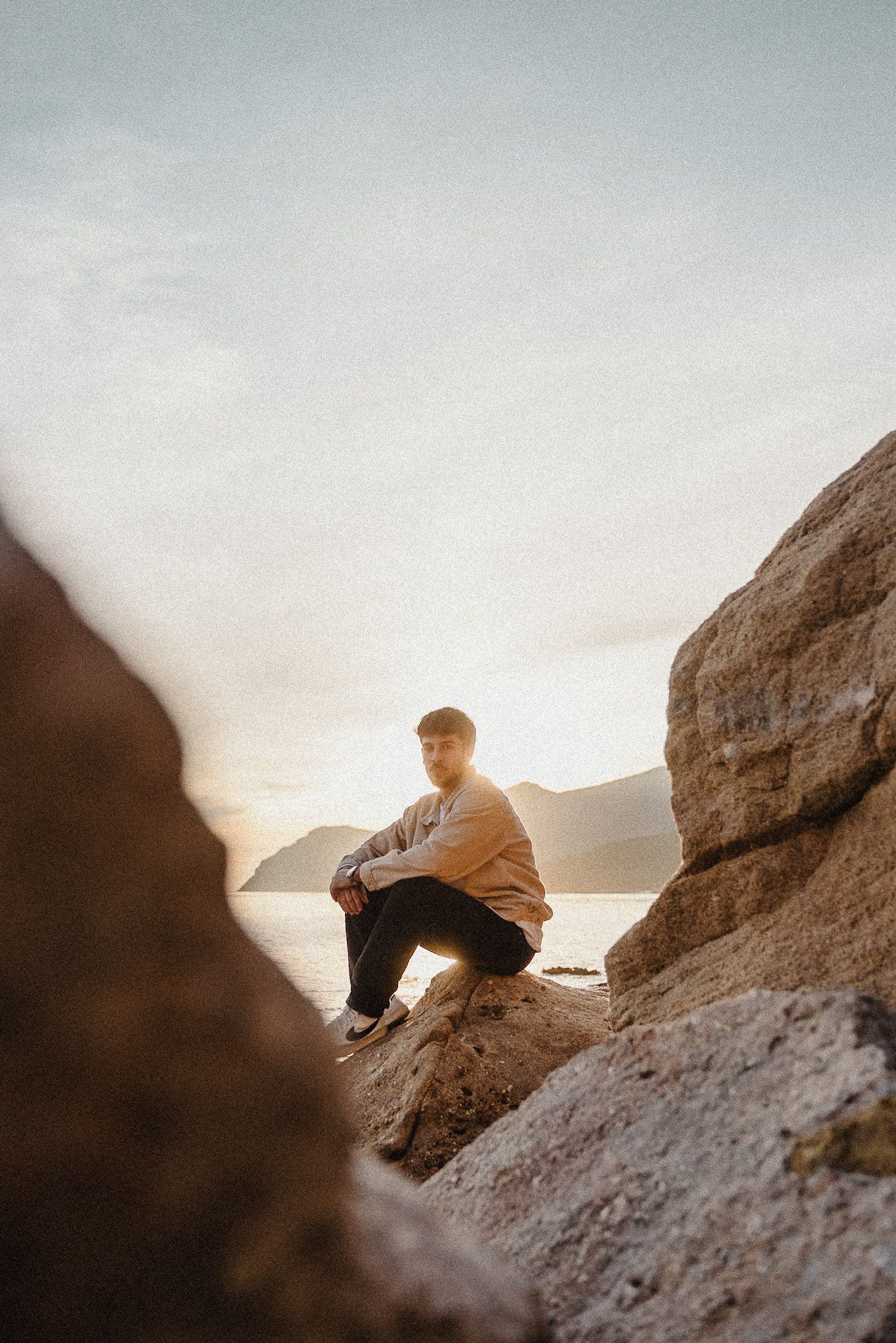 Person sitting on coastal rocks at sunset overlooking mountain views.