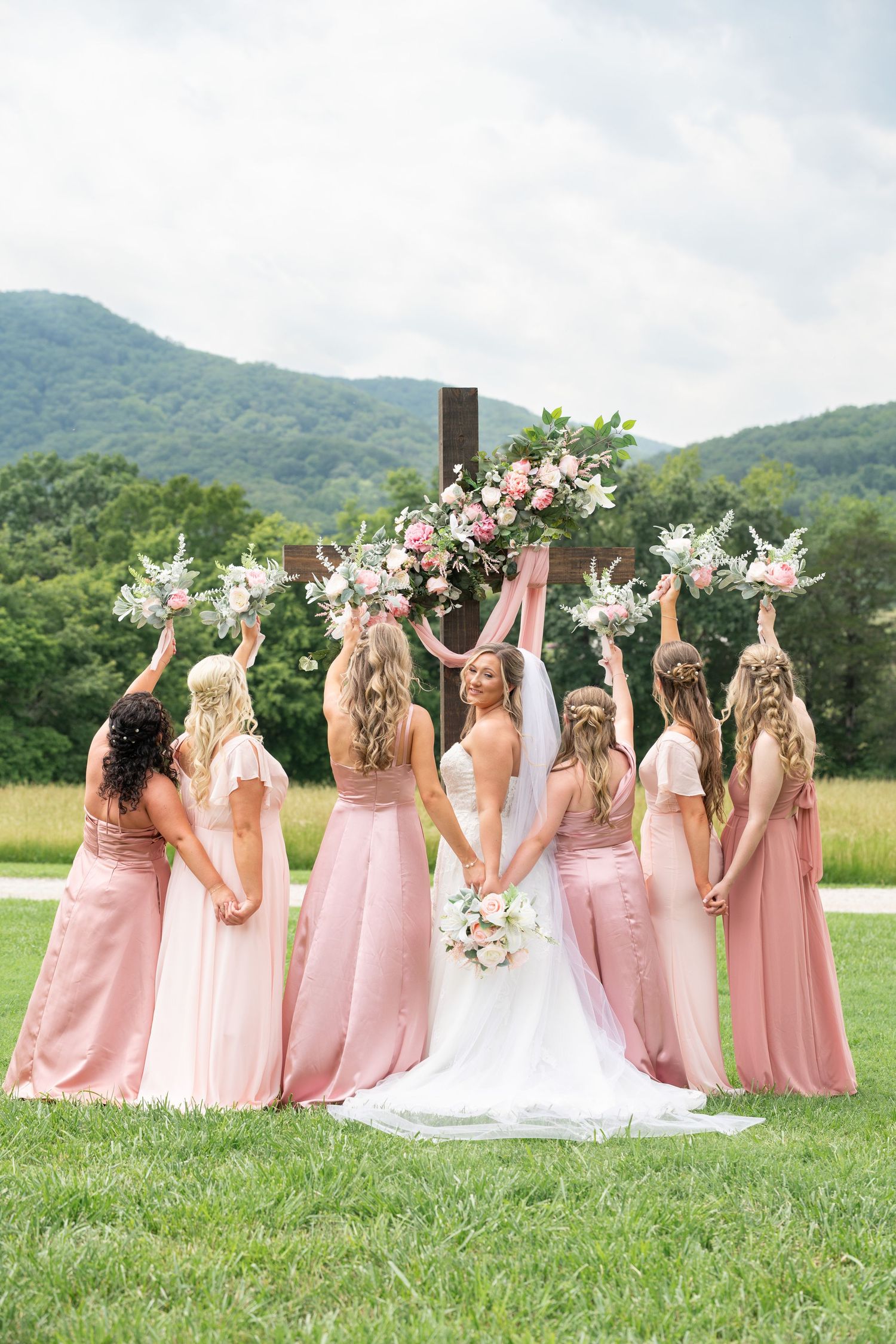 Bride and bridesmaids in pink dresses raise bouquets before a floral cross with mountain views.