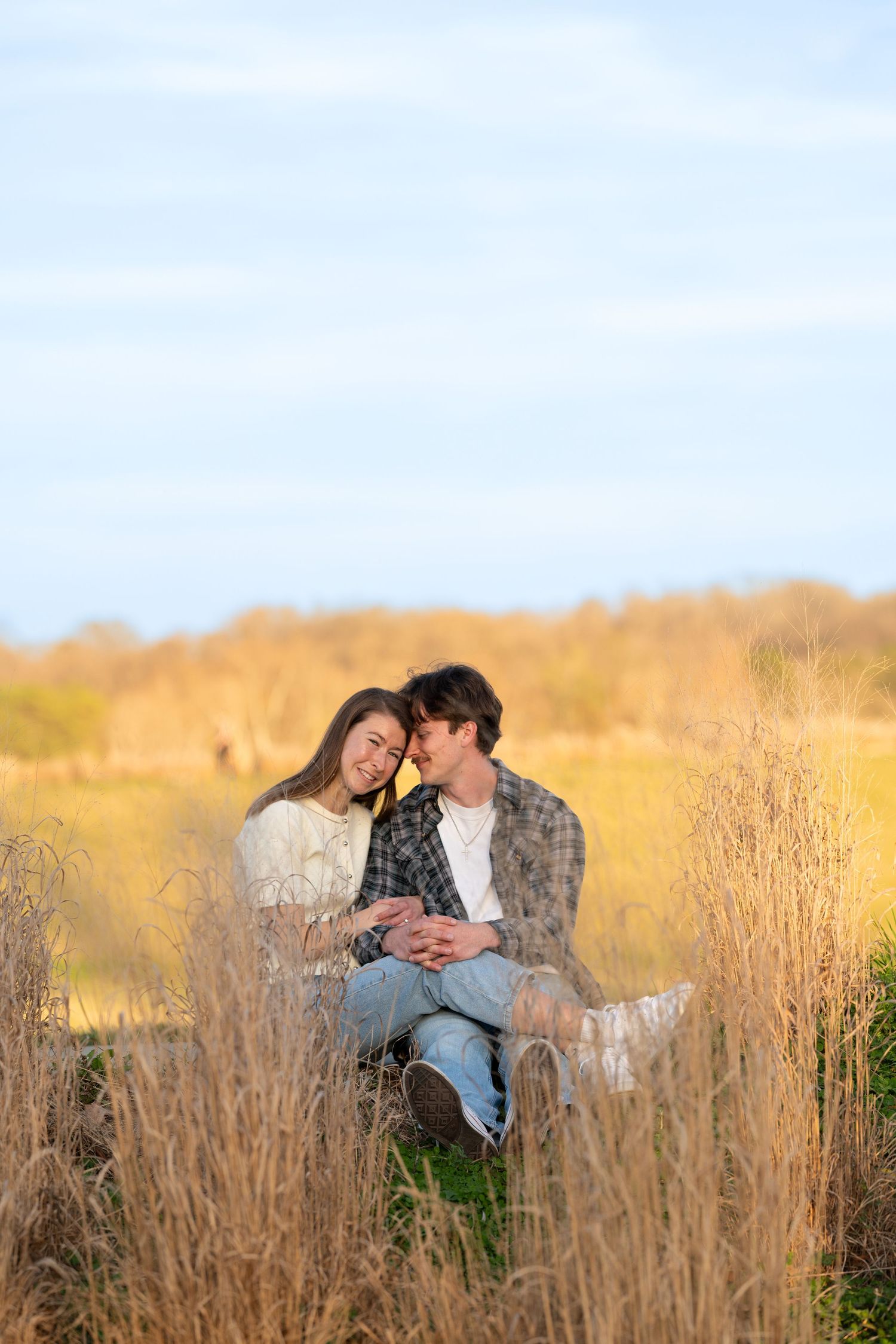 Romantic couple sitting together in golden autumn field grass, sharing a tender moment under a blue sky.