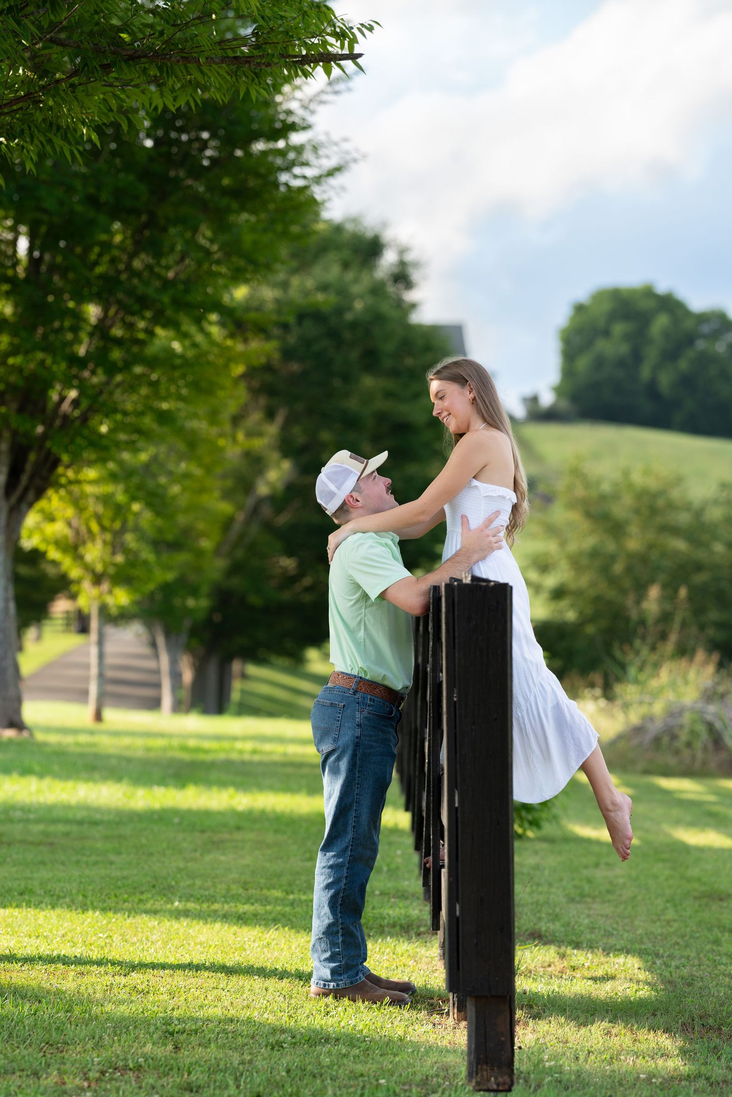 A man in a cowboy hat lifts a woman in a white dress outdoors on a sunny green lawn.