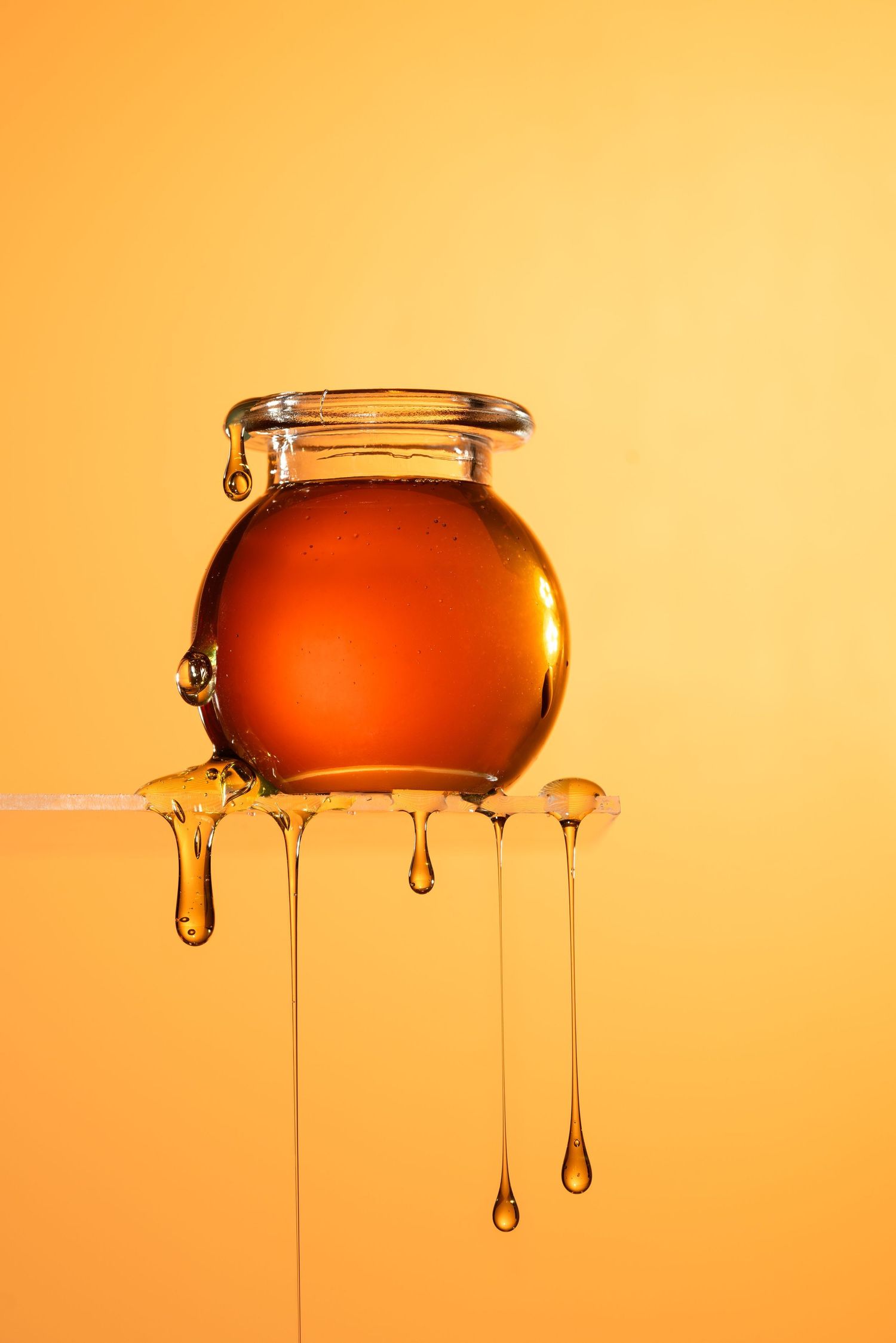 A glass jar filled with golden honey sitting on a wooden stick with honey dripping down on orange background.