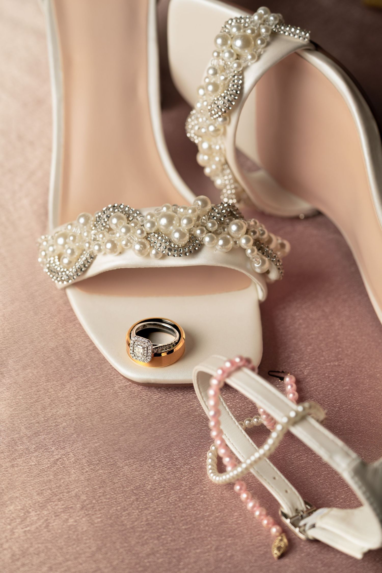 Elegant white bridal heels with pearl embellishments and wedding rings displayed on a soft pink background.
