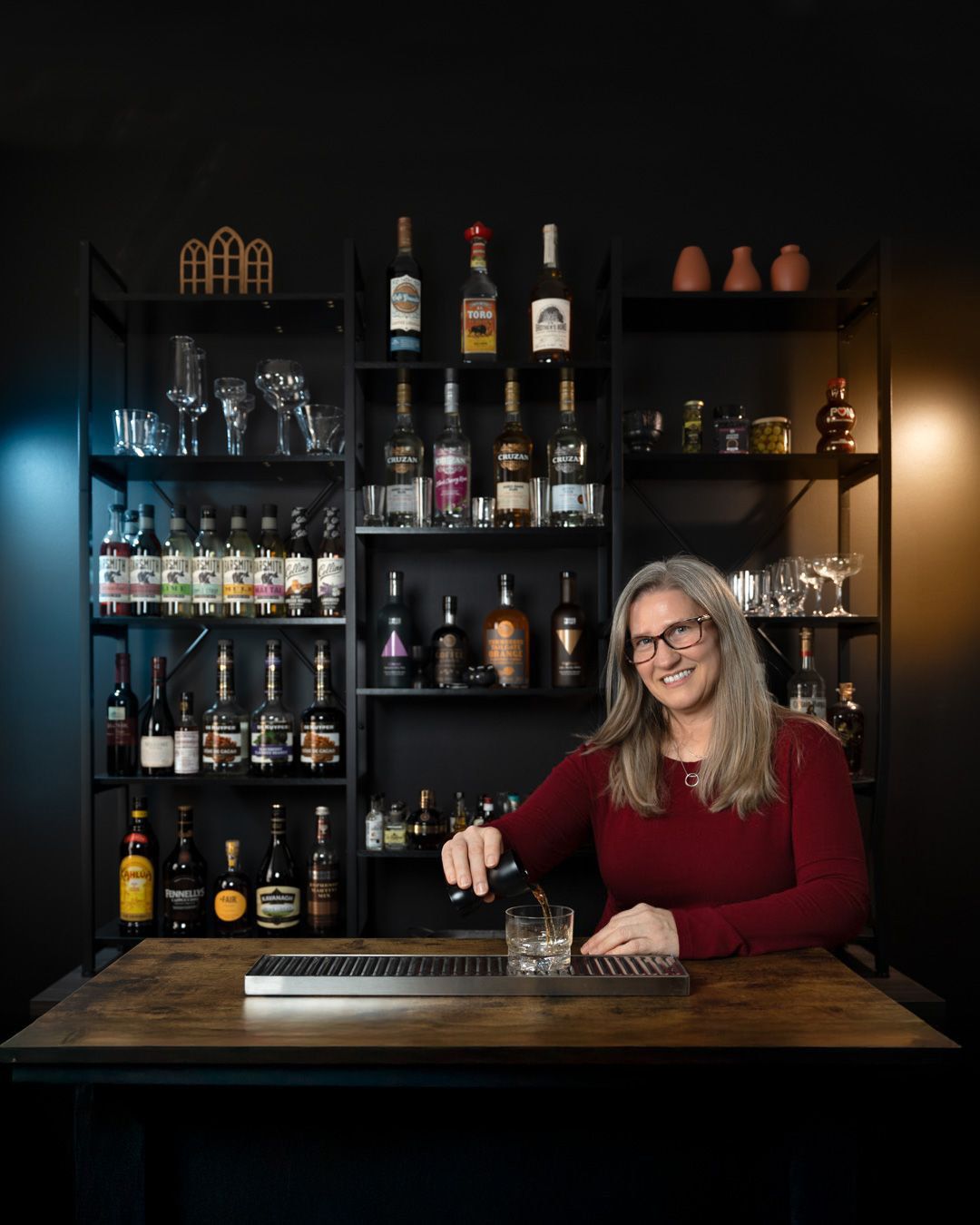 Woman bartender smiling behind a well-stocked bar shelf, preparing a cocktail in a cozy dark-themed lounge.