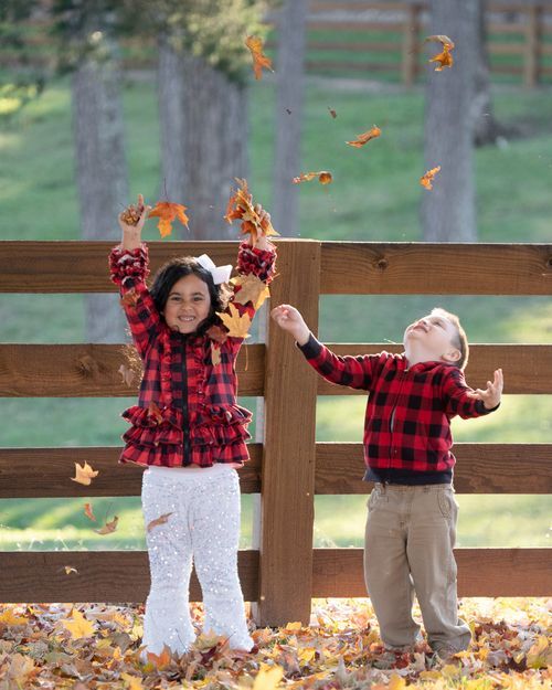 Two children in matching red plaid shirts toss autumn leaves in the air near a wooden fence.