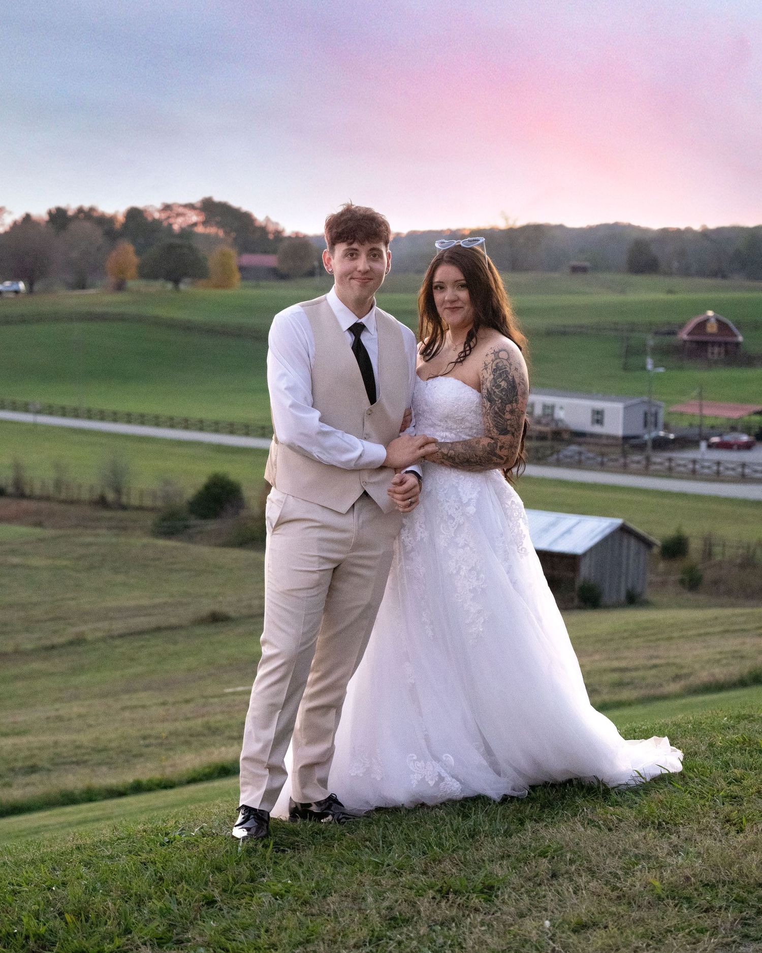 Bride and groom posing at sunset on a green hillside farm, she in white gown, he in tan suit with black tie.