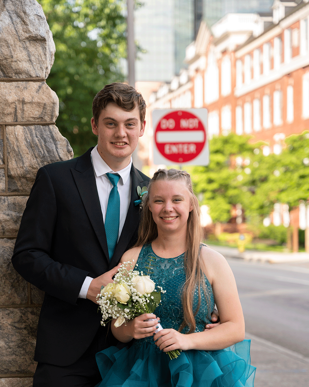 A young couple in teal formal wear poses on a city street, the woman holding a white bouquet near a Do Not Enter sign.