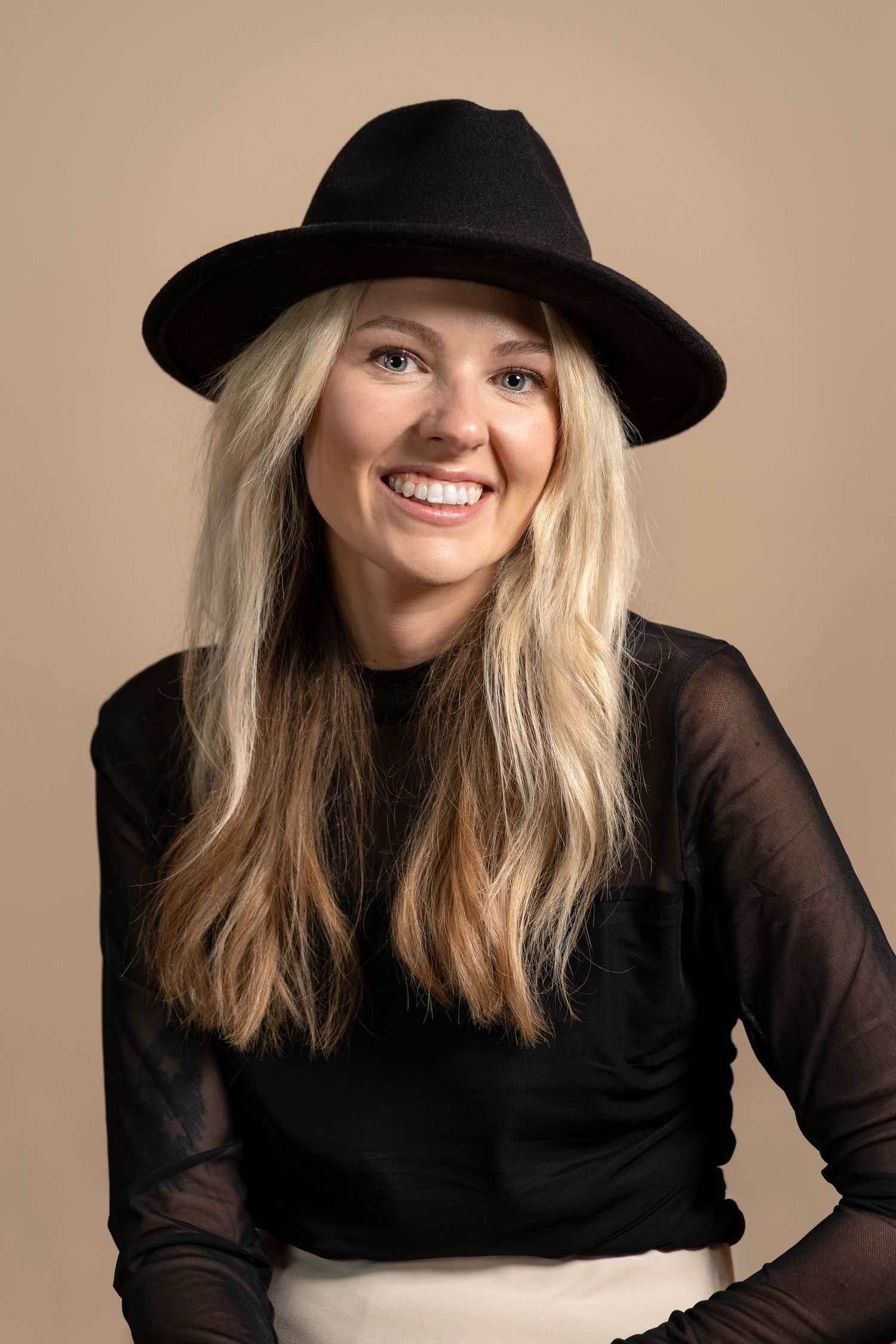 Smiling blonde woman wearing a black wide-brim hat and sheer black top, posing against a beige background.