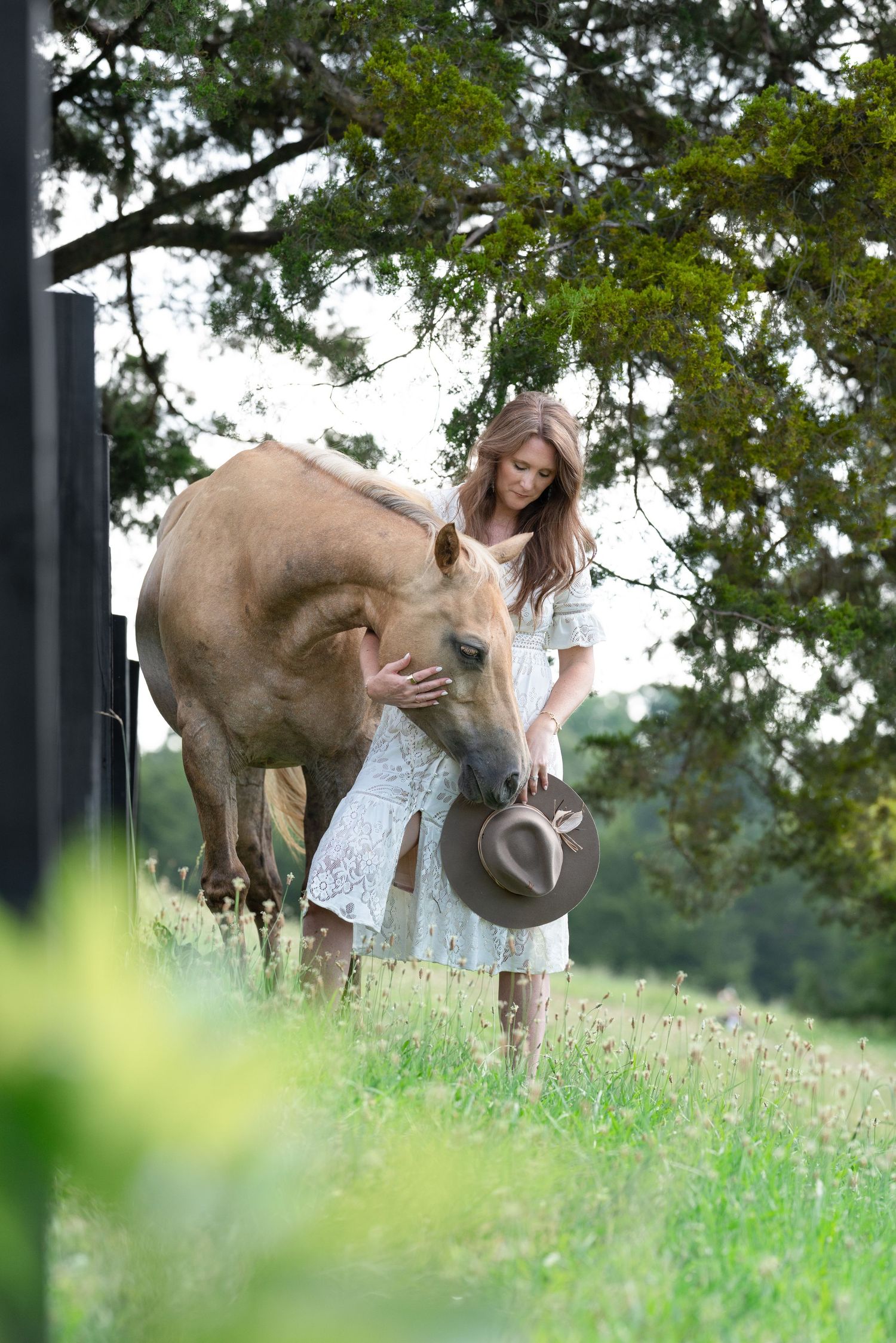 Young woman in white dress holds cowboy hat while gently petting a palomino horse in a lush green field.
