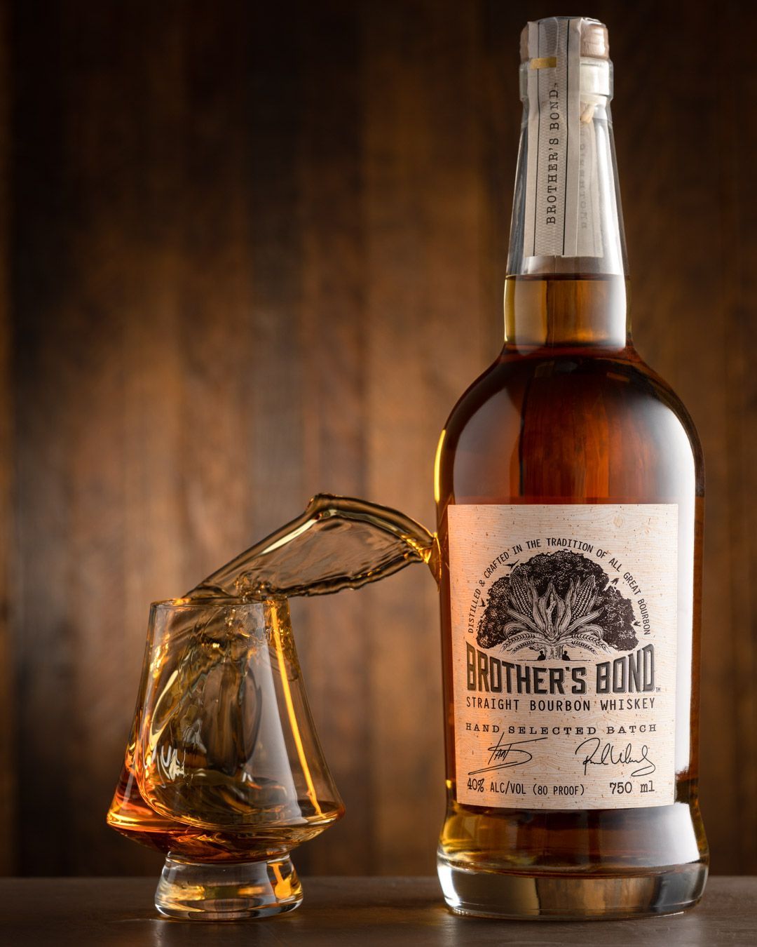 Brother's Bond Straight Bourbon Whiskey bottle beside a tilted crystal glass on a dark wooden surface.