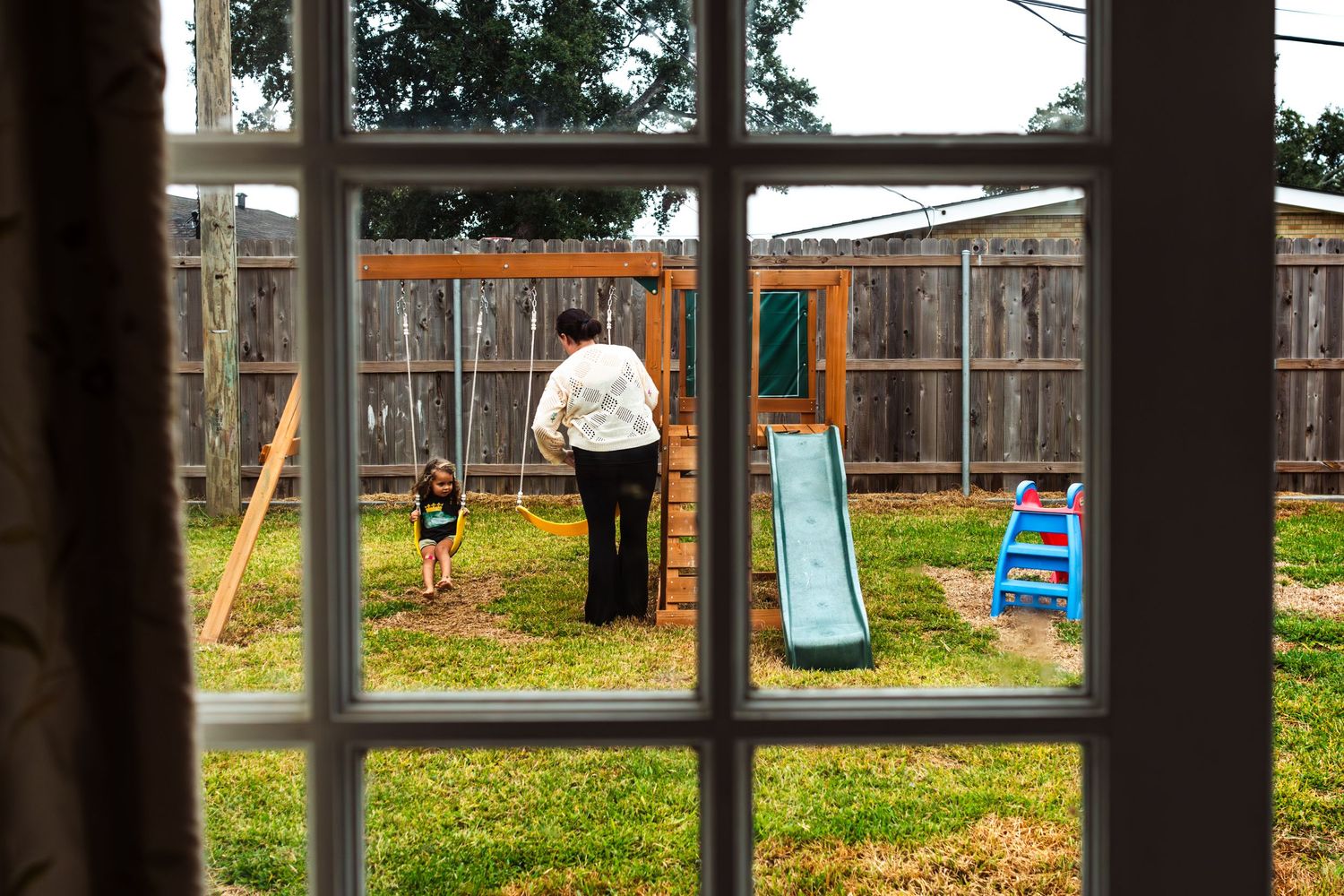 Wooden playground equipment including swings and climbing structures on a grassy hill in a residential backyard.
