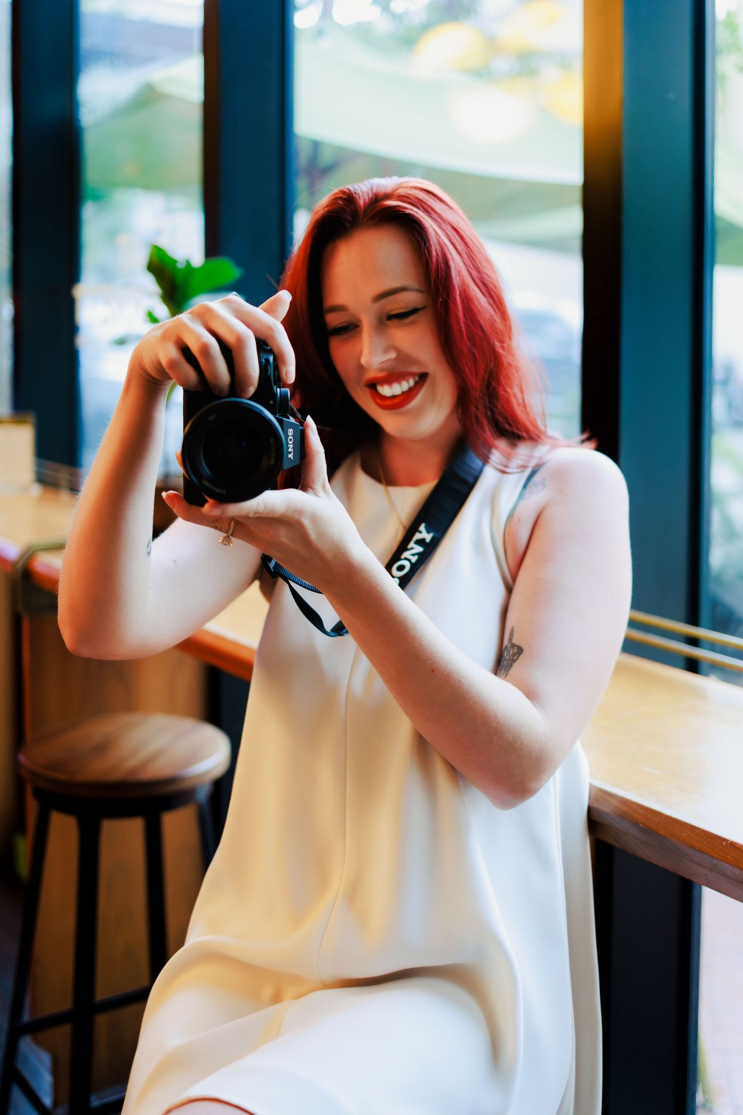 Smiling redhead woman in white dress holding a Sony camera, sitting in a bright cafe interior.