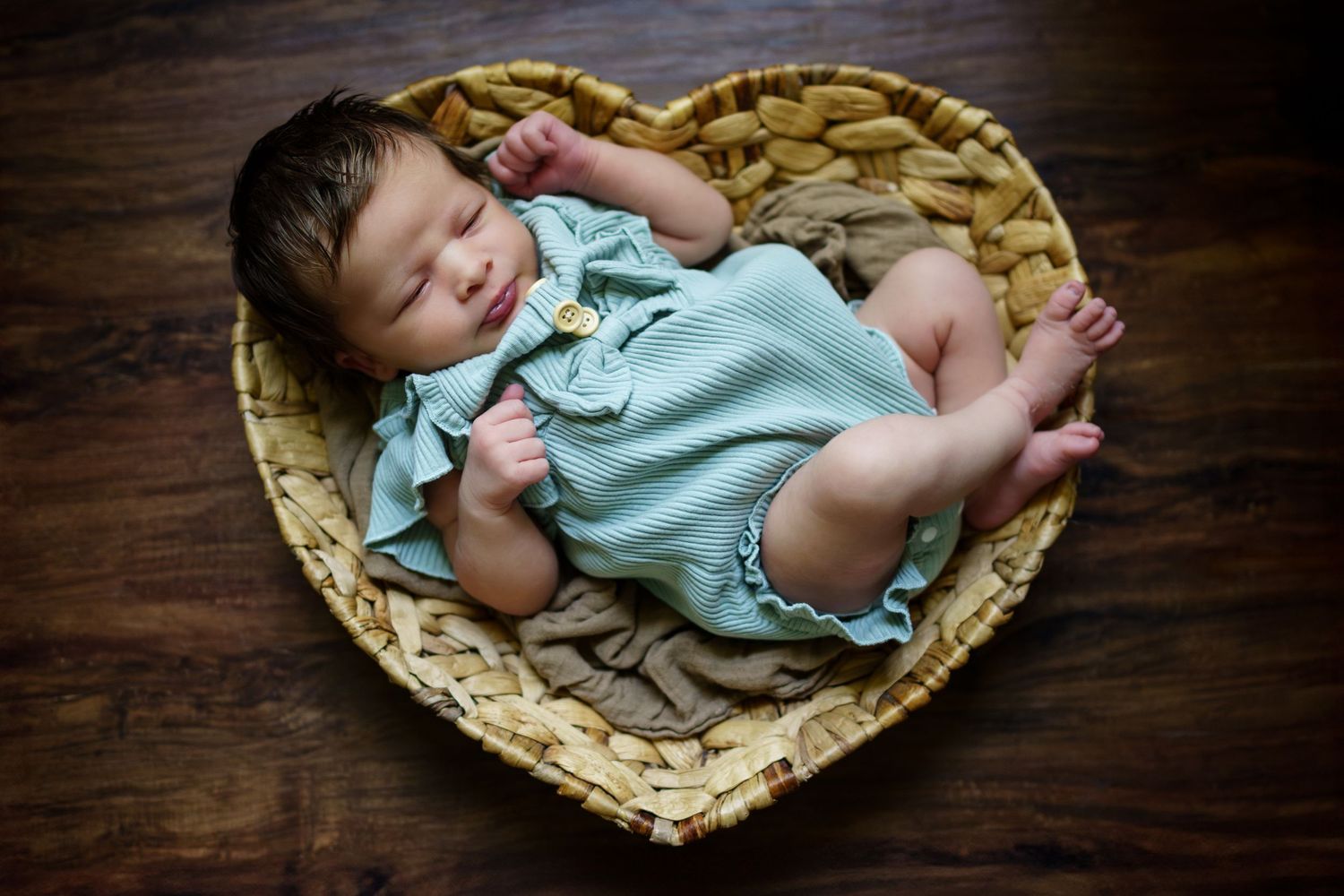 A sleeping baby wearing mint green romper rests peacefully in a heart-shaped woven basket.