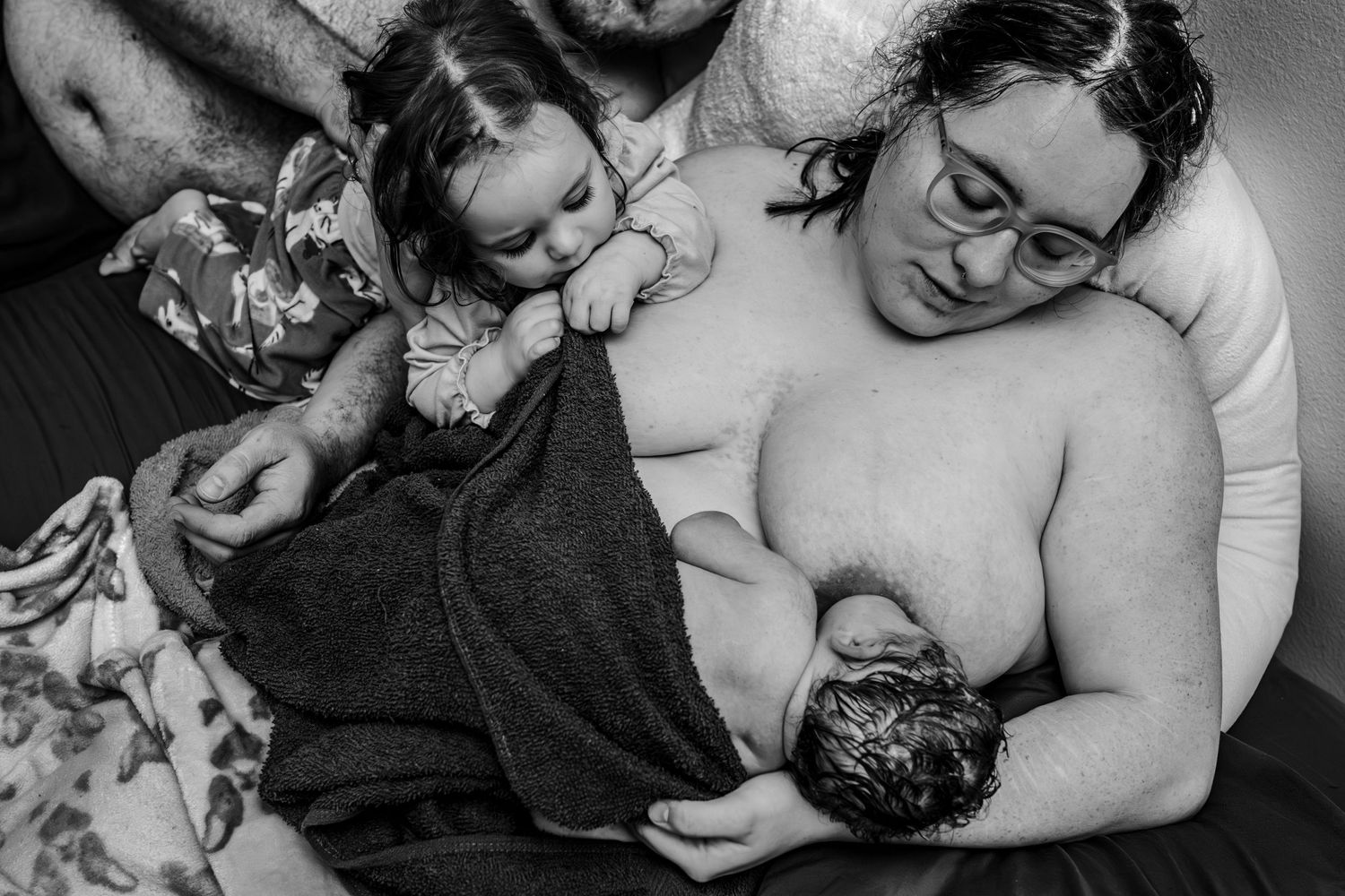 Black and white intimate photograph showing a mother breastfeeding her newborn baby while another child looks on.