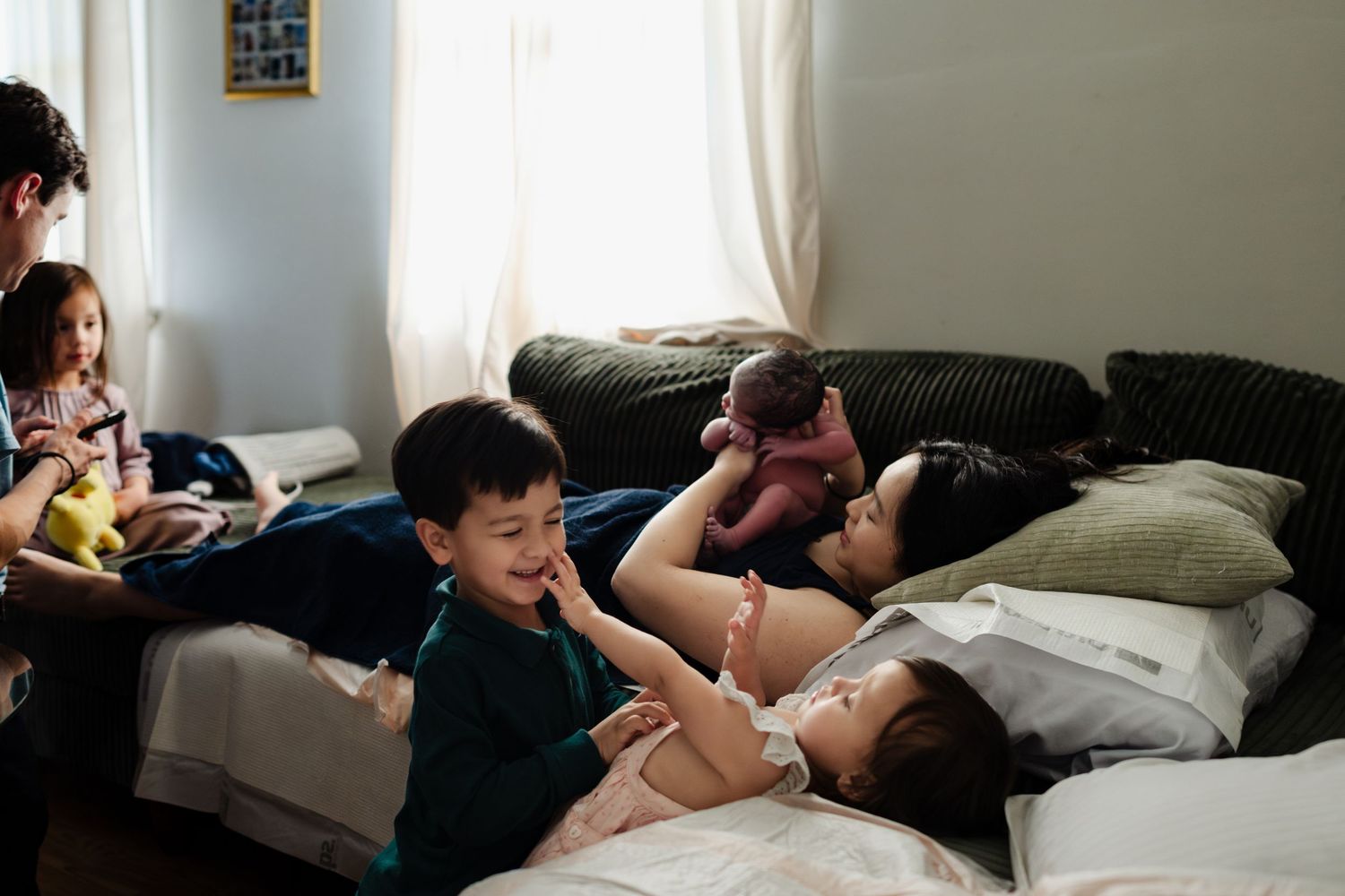 A mother rests in bed with her newborn baby surrounded by her young children in a cozy bedroom.