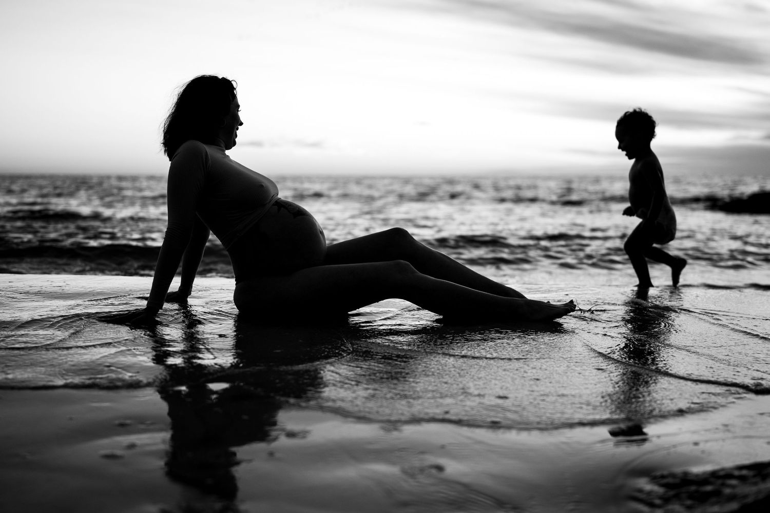 Silhouette of pregnant woman and child on beach at sunset.
