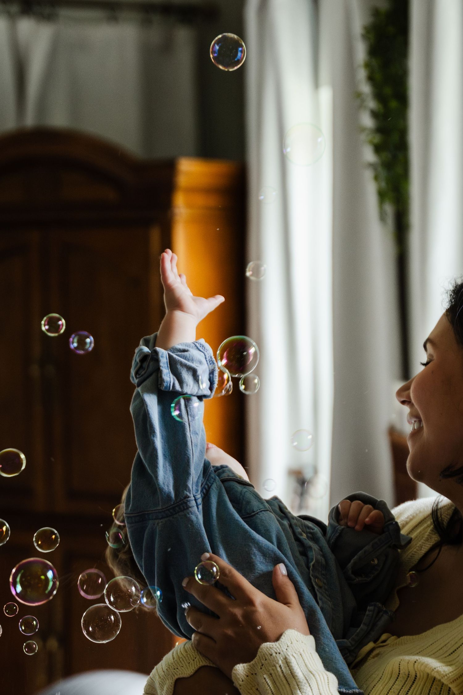 Parent and child playing with soap bubbles by a window in casual denim clothing during a joyful indoor moment.