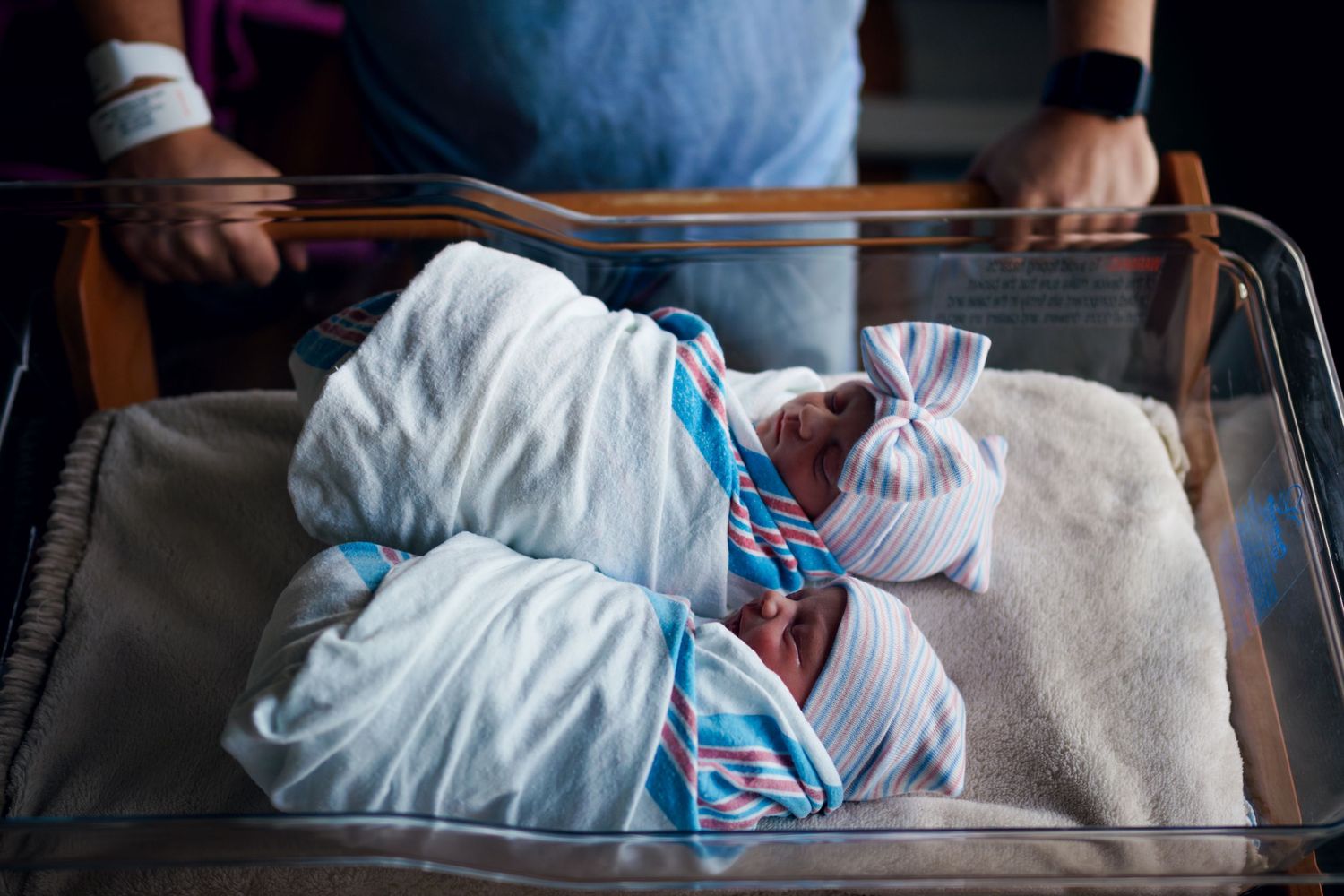 Two newborn twins swaddled in white blankets with colorful hats resting in a hospital bassinet.