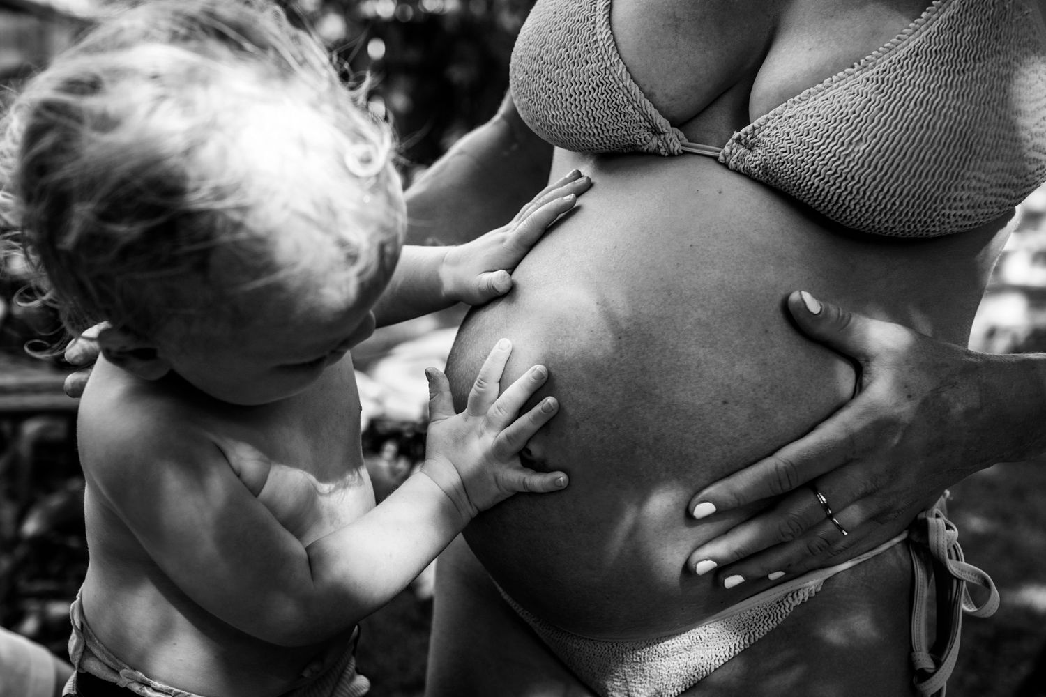 Intimate black and white photograph of a pregnant belly being touched by small hands in tender curiosity.