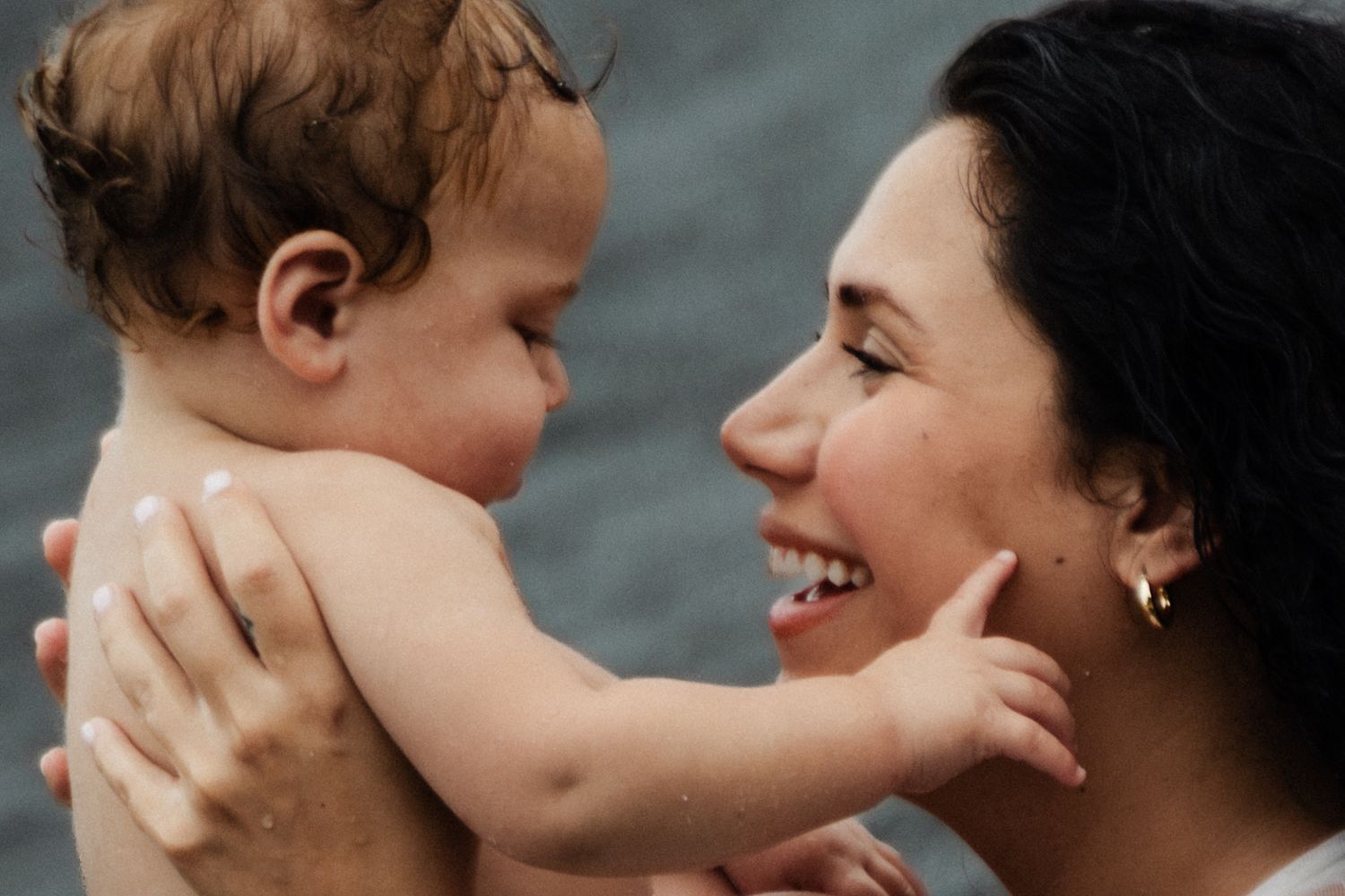 A joyful moment shared between a parent and baby as they smile and touch each other's faces against a gray background.