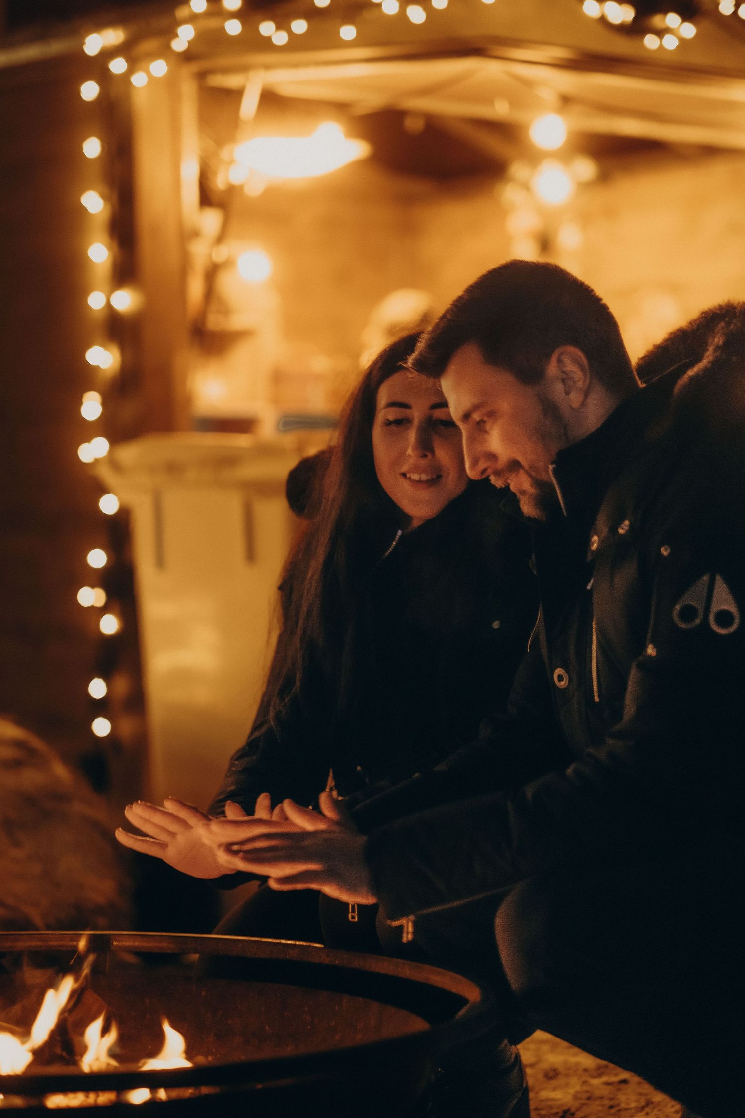 couple warming up their hands over fire with Christmas lights behind them while being captured by cinematic couple photographer in Stockholm