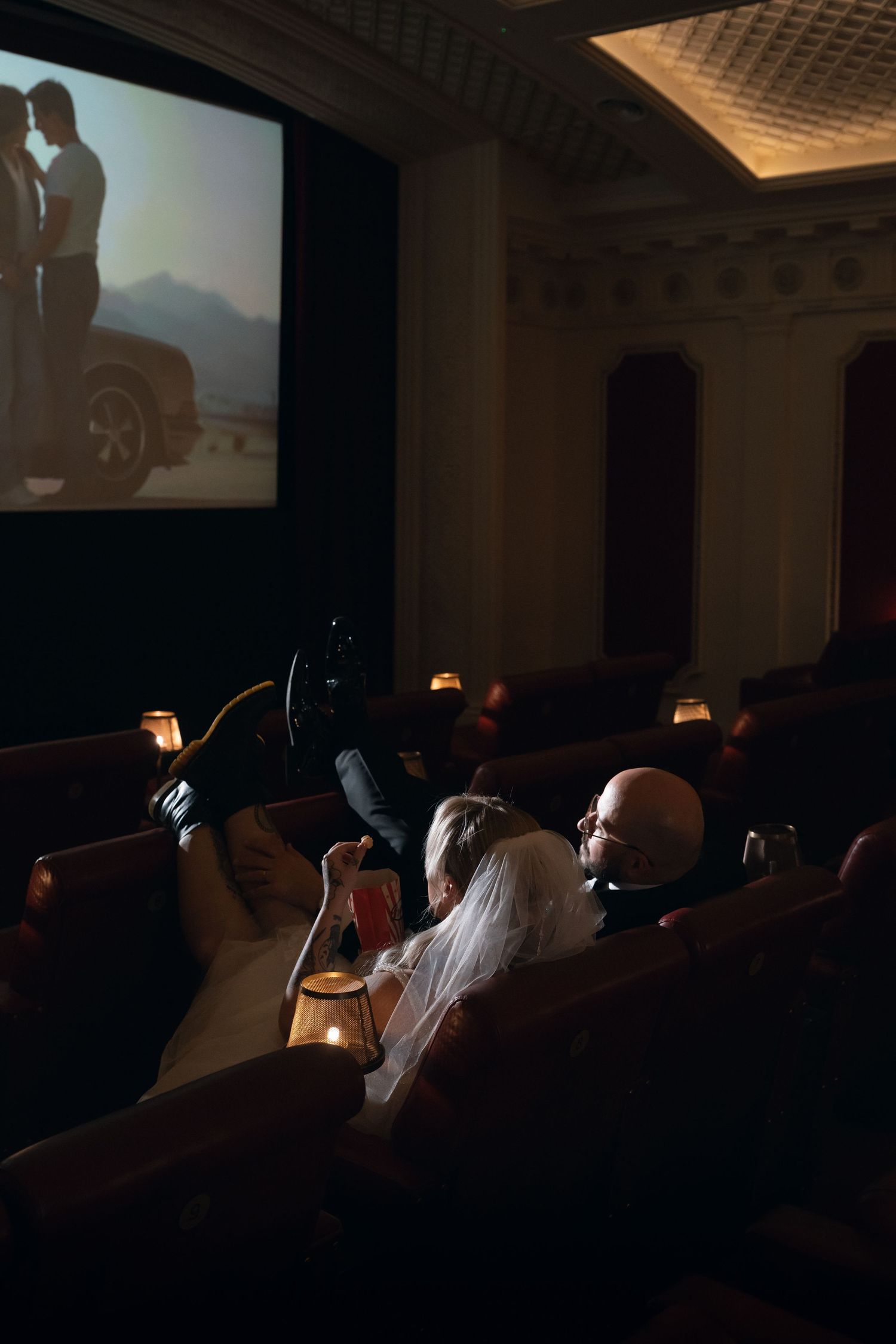 Bride and groom watching a romantic film in a luxurious candlelit cinema with plush seating.