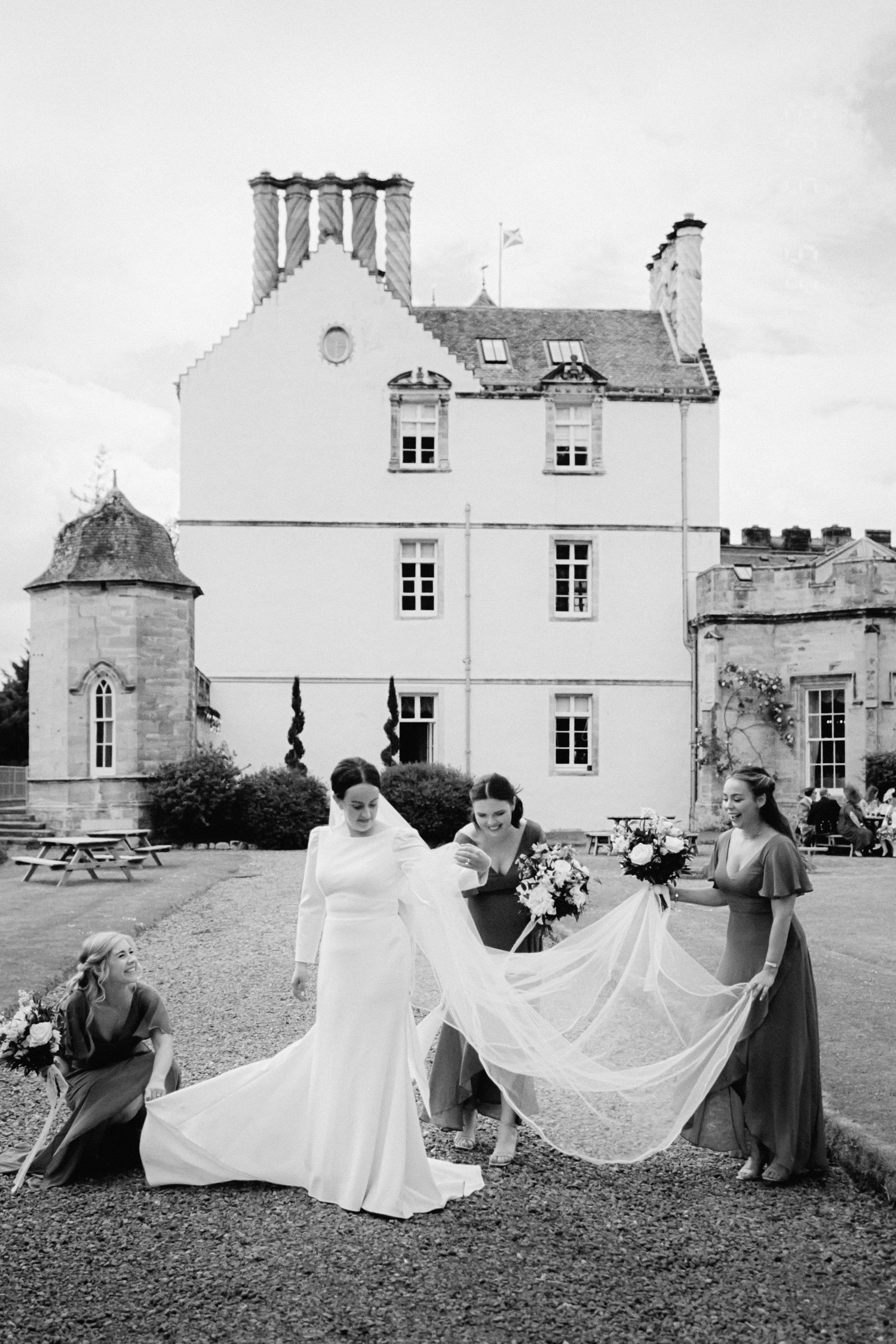 A series of four black and white photographs showing a wedding dress being arranged on a manor house lawn.