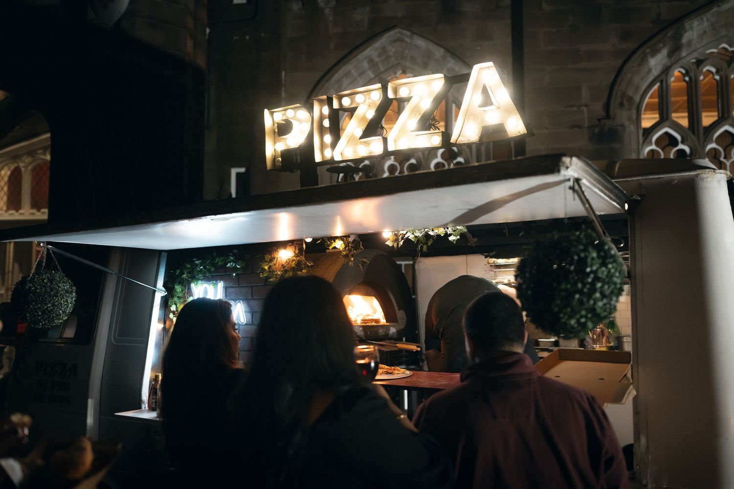 Illuminated pizza restaurant sign glows in the dark against a night sky with outdoor seating area visible.