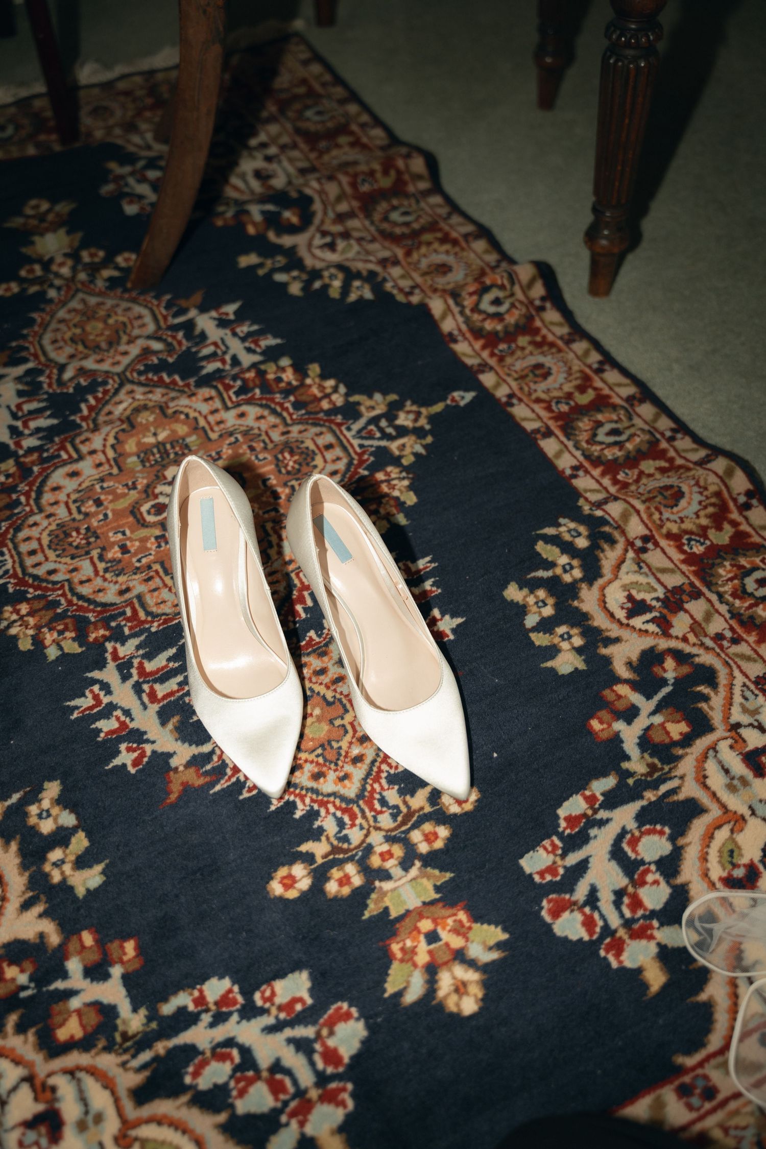 A pair of elegant white bridal heels placed on a dark vintage carpet with ornate floral patterns.