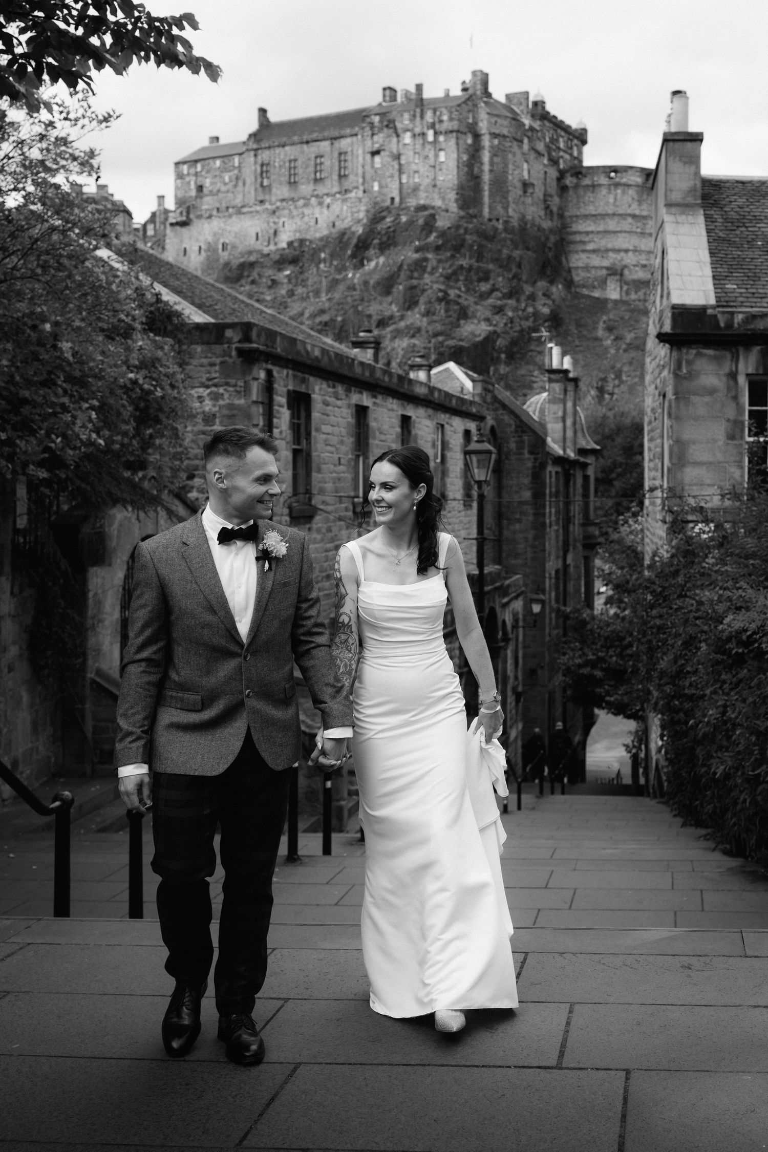 A couple walks along a historic cobblestone path with Edinburgh Castle looming in the background on their wedding day.