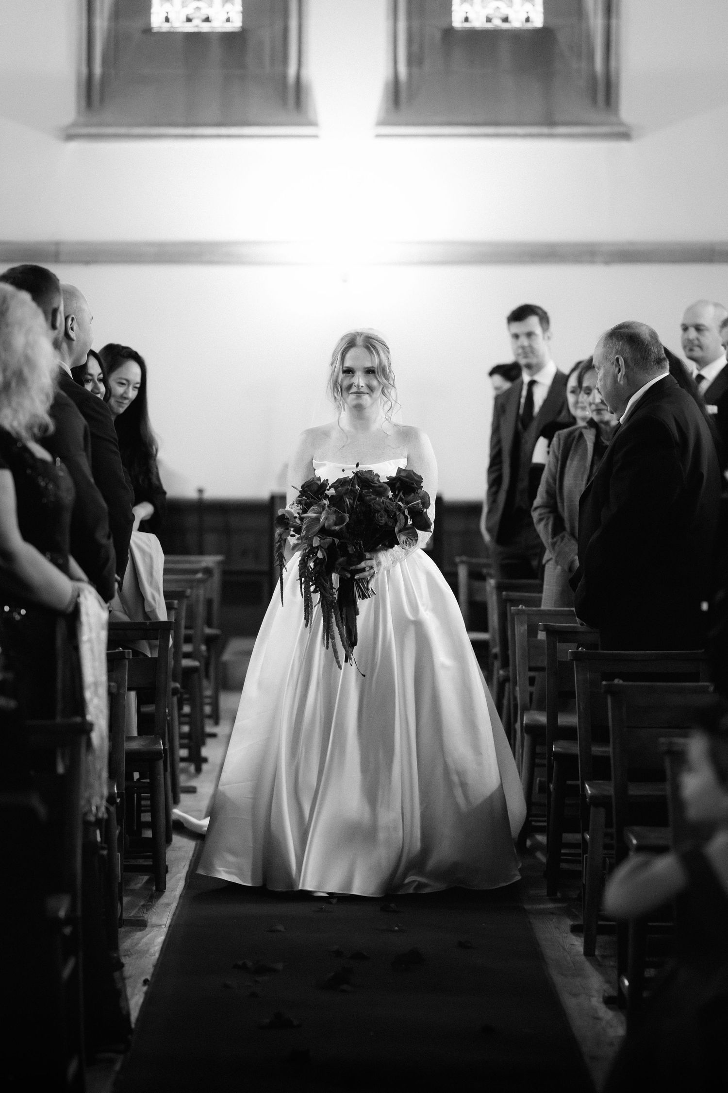 A black and white wedding photograph showing a bride in a classic ballgown walking down the church aisle.