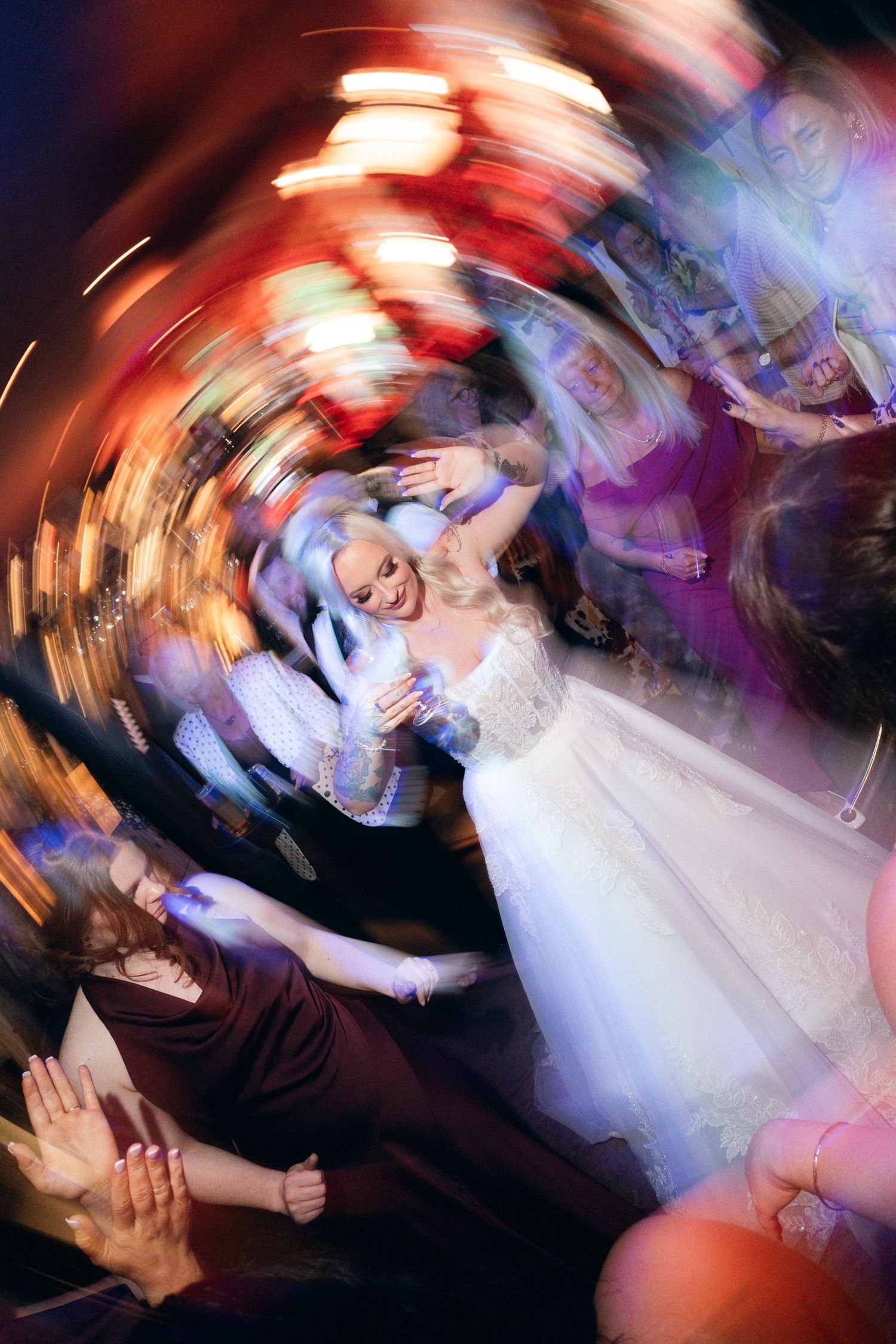 Bride in white gown dancing joyfully with guests at a wedding reception, captured with a swirling motion blur effect.