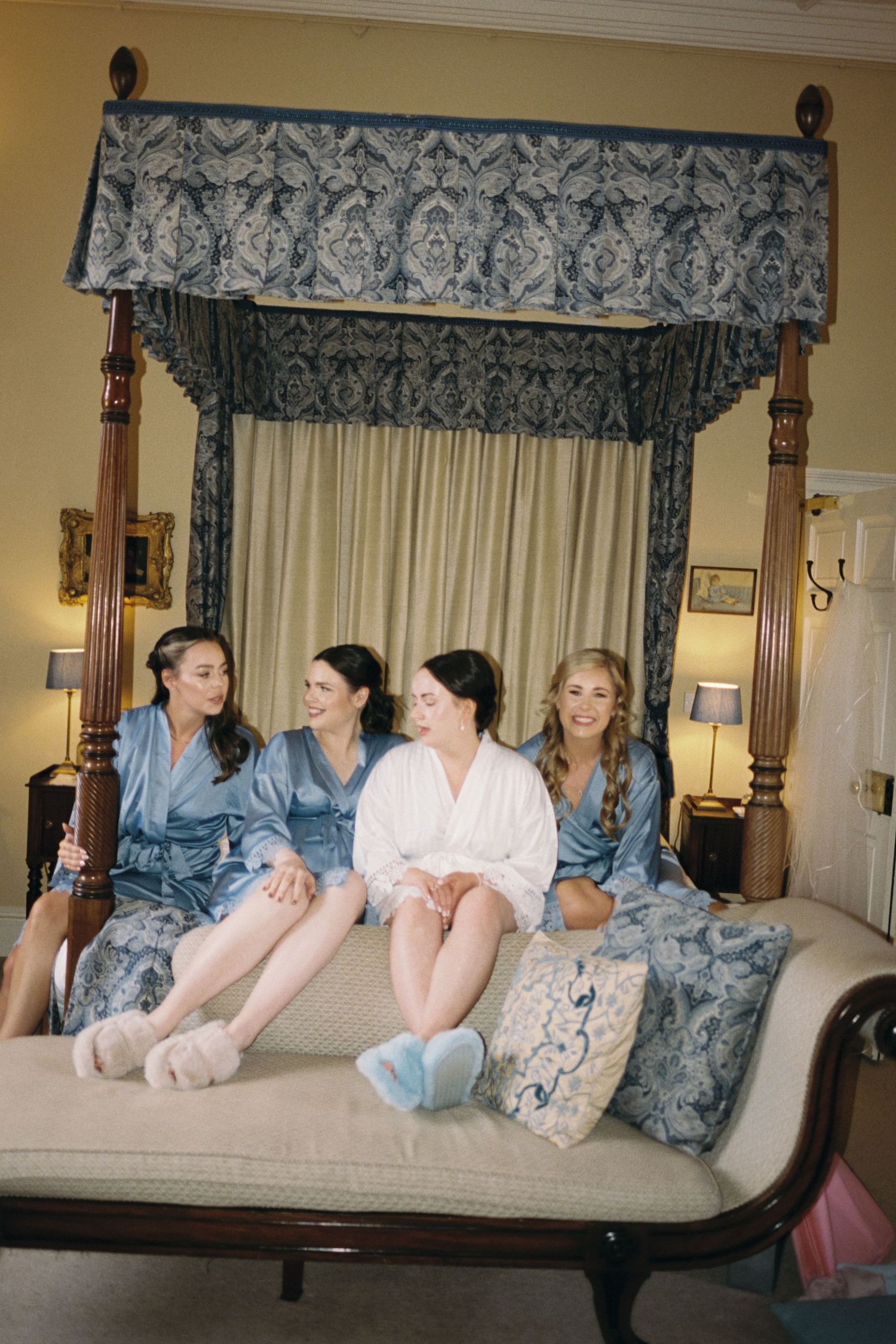 A group in matching silk robes sits together on an elegant canopy bed with decorative damask curtains.